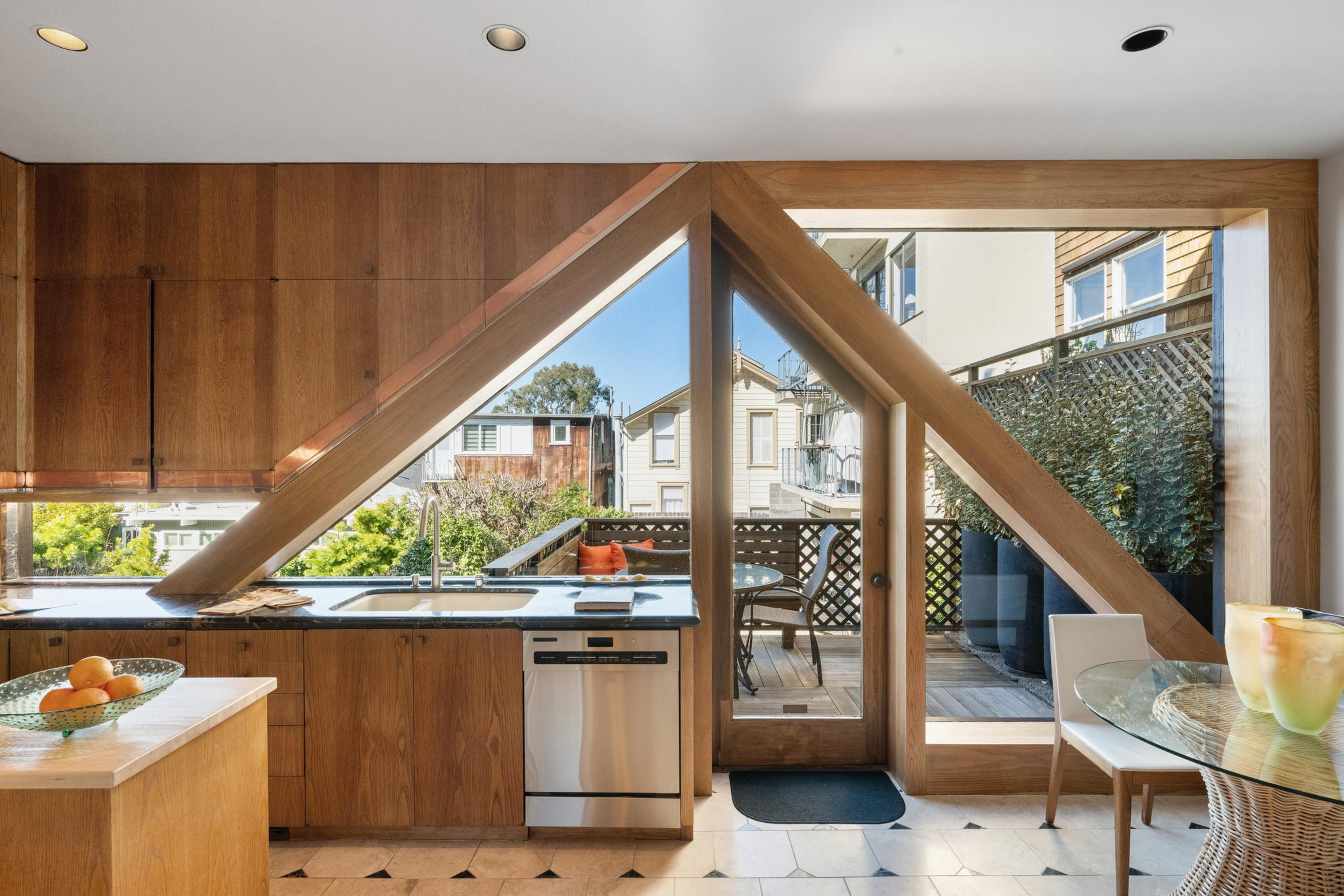 A kitchen with wooden cabinets and an angled window revealing a patio. The interior includes a glass table, chairs, and a bowl of fruit on the countertop.