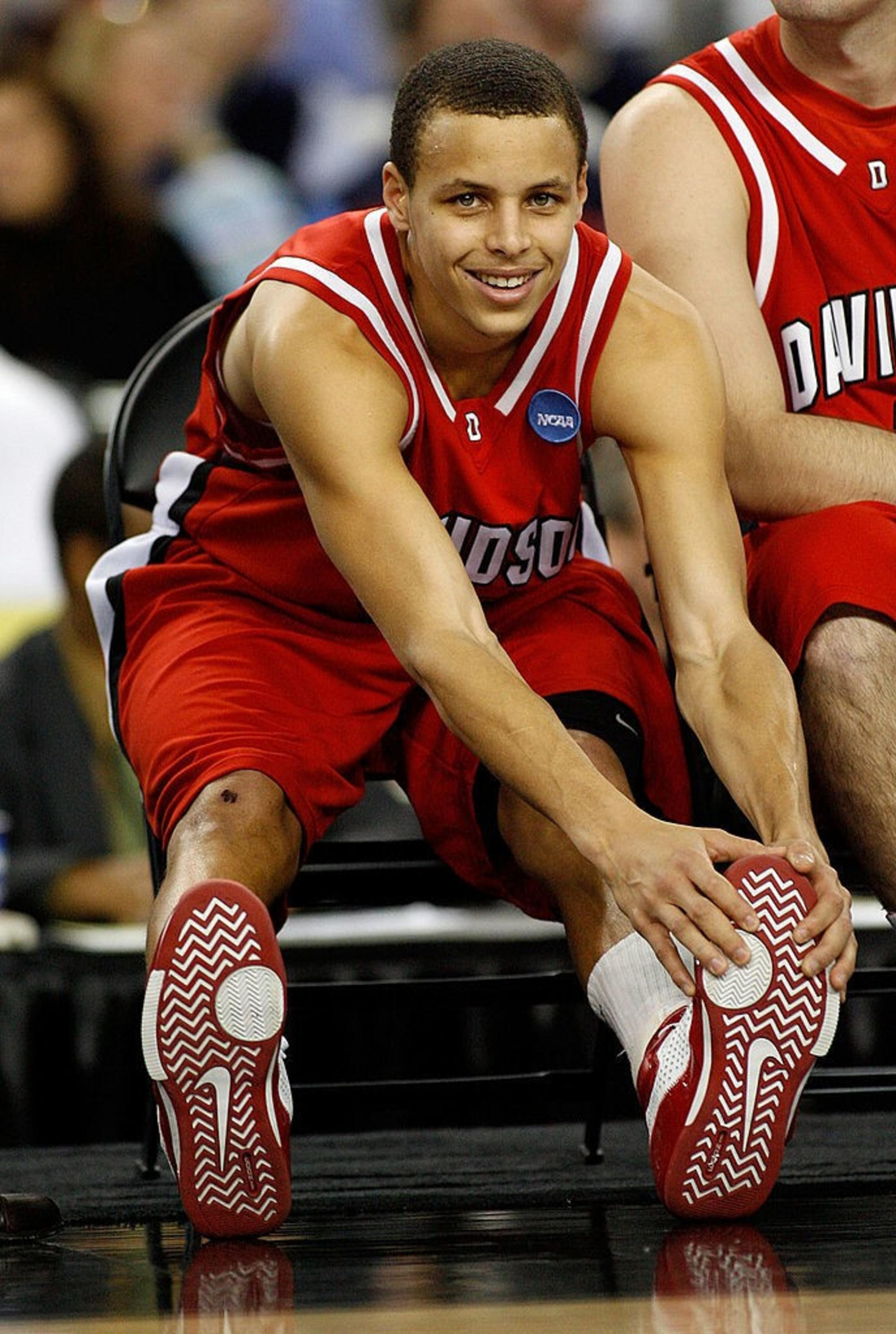 A basketball player in a red Davidson uniform is sitting down, stretching forward to touch his shoes, smiling, with a crowd blurred in the background.