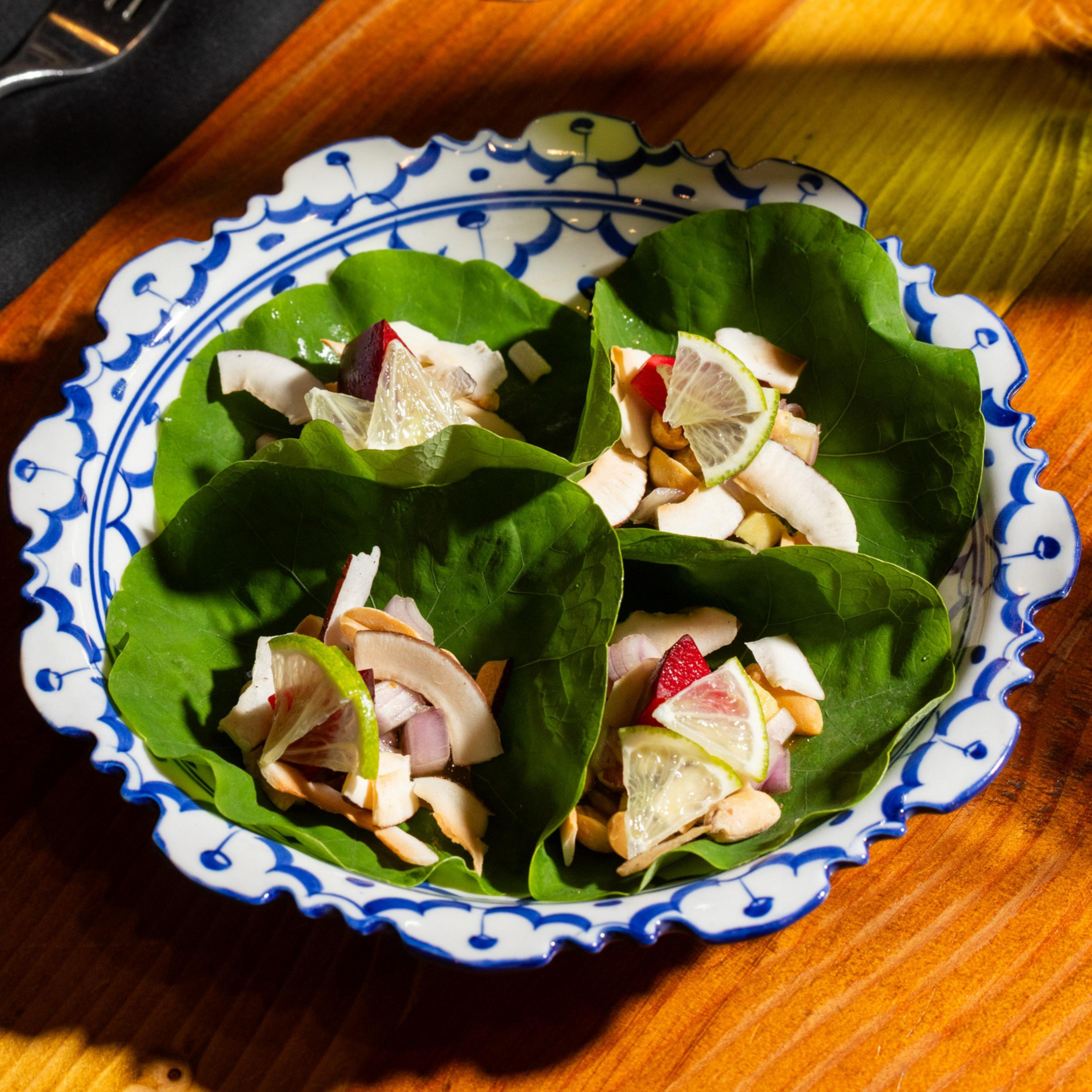 Four green leaves on a decorative plate, each holding chopped ingredients like coconut, lime, and onions, arranged neatly with sunlight streaming on them.