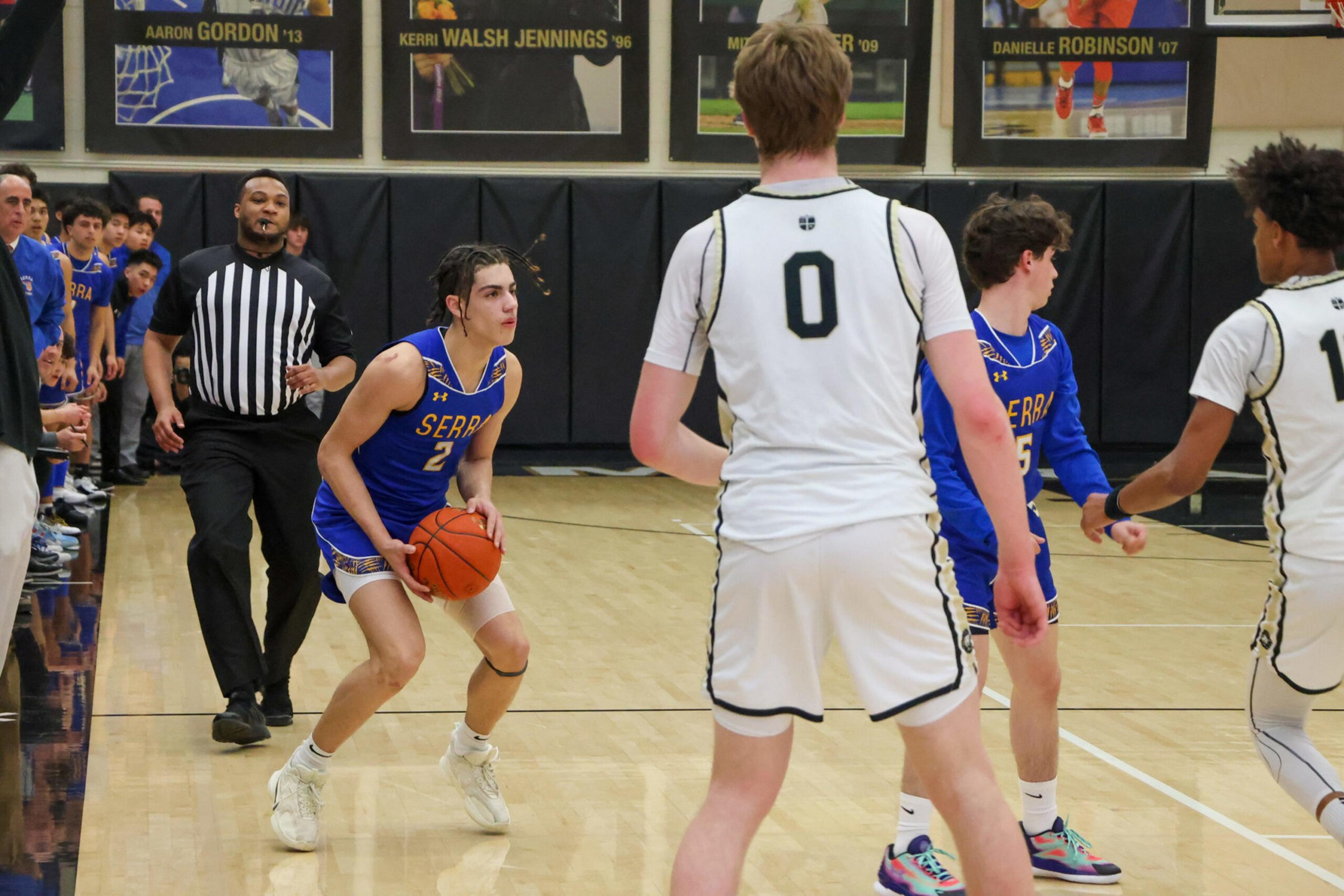 A basketball player in a blue jersey prepares to shoot while guarded by two players in white jerseys, with a referee and spectators in the background.
