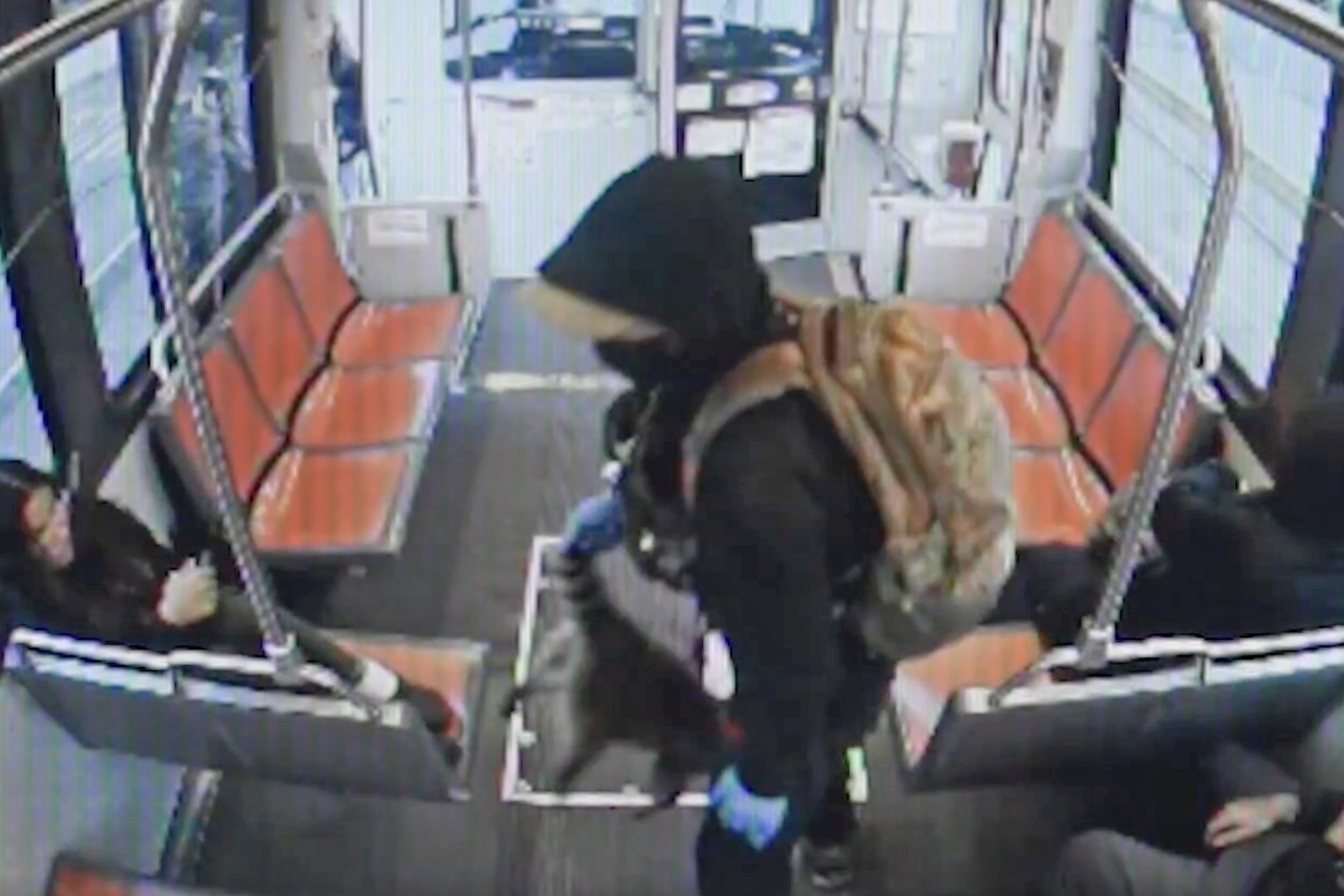 A person with a backpack and gloves enters a bus, wearing a hoodie and mask. Two passengers sit on orange seats, one on each side, with empty seats visible.