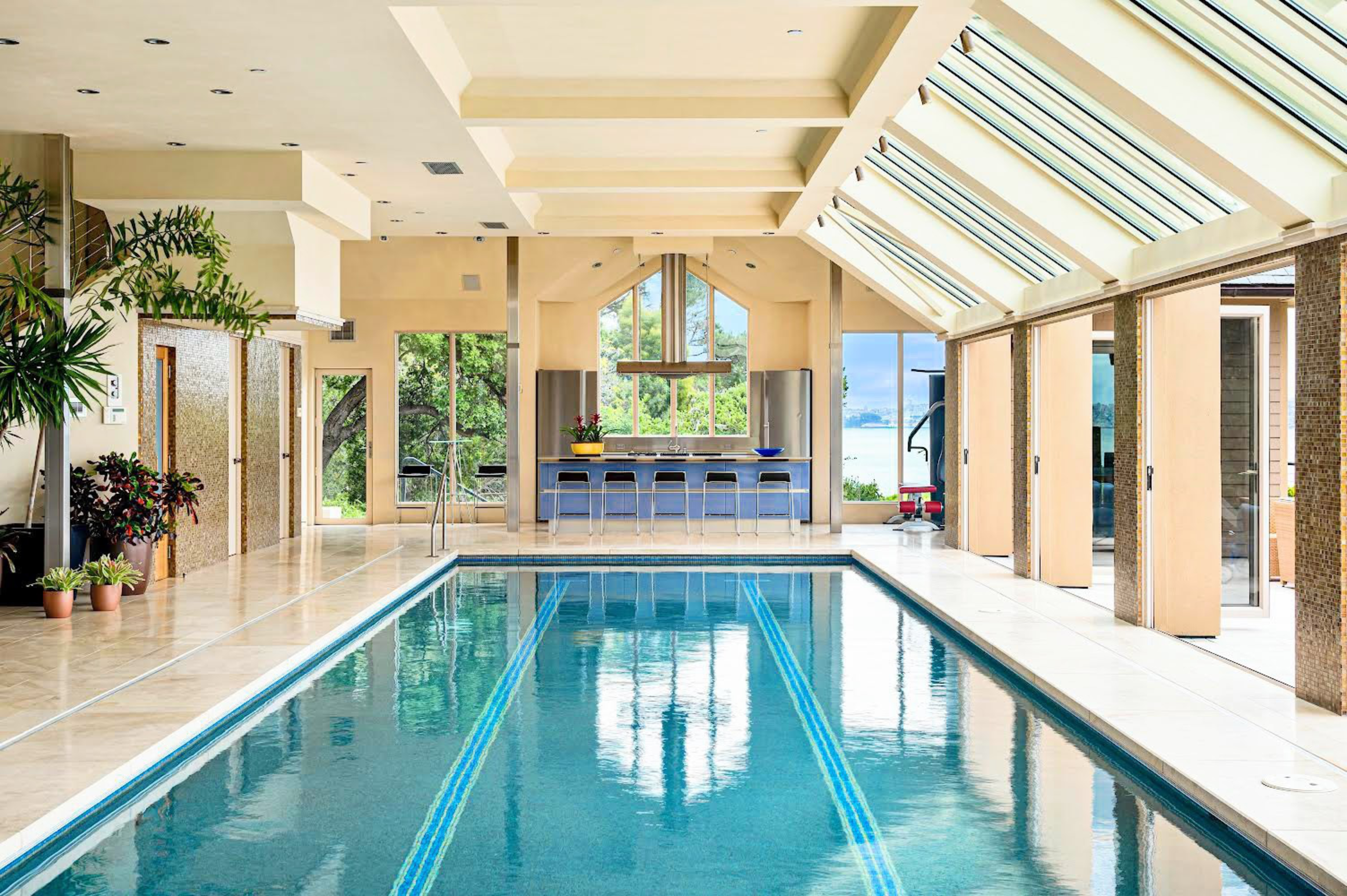 An indoor swimming pool with tiled flooring is surrounded by large windows and skylights, with a kitchen area and bar stools at one end.