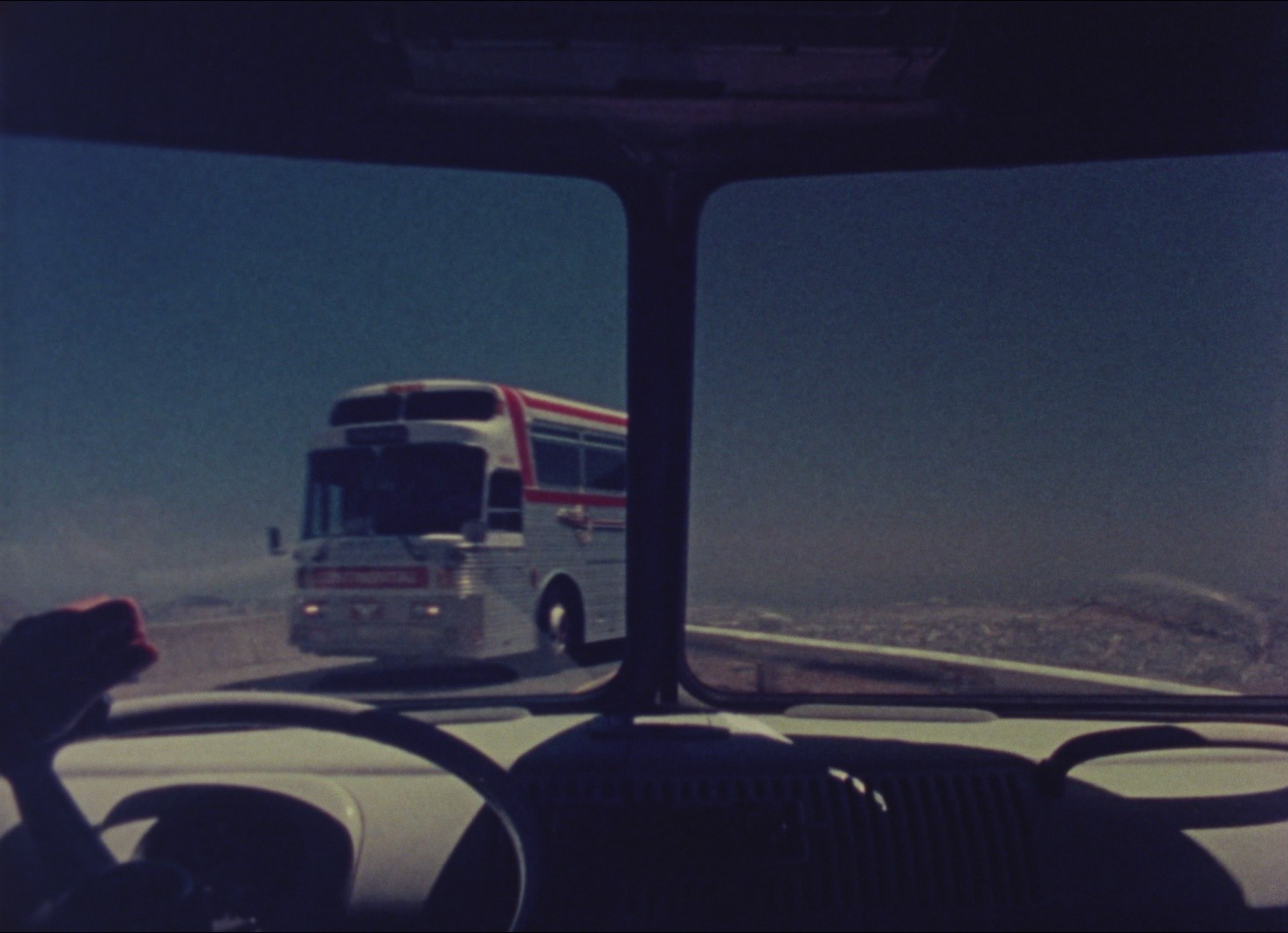A bus is approaching on a winding road as seen through a vehicle's windshield. The driver’s hand grips the steering wheel, and a clear sky forms the backdrop.