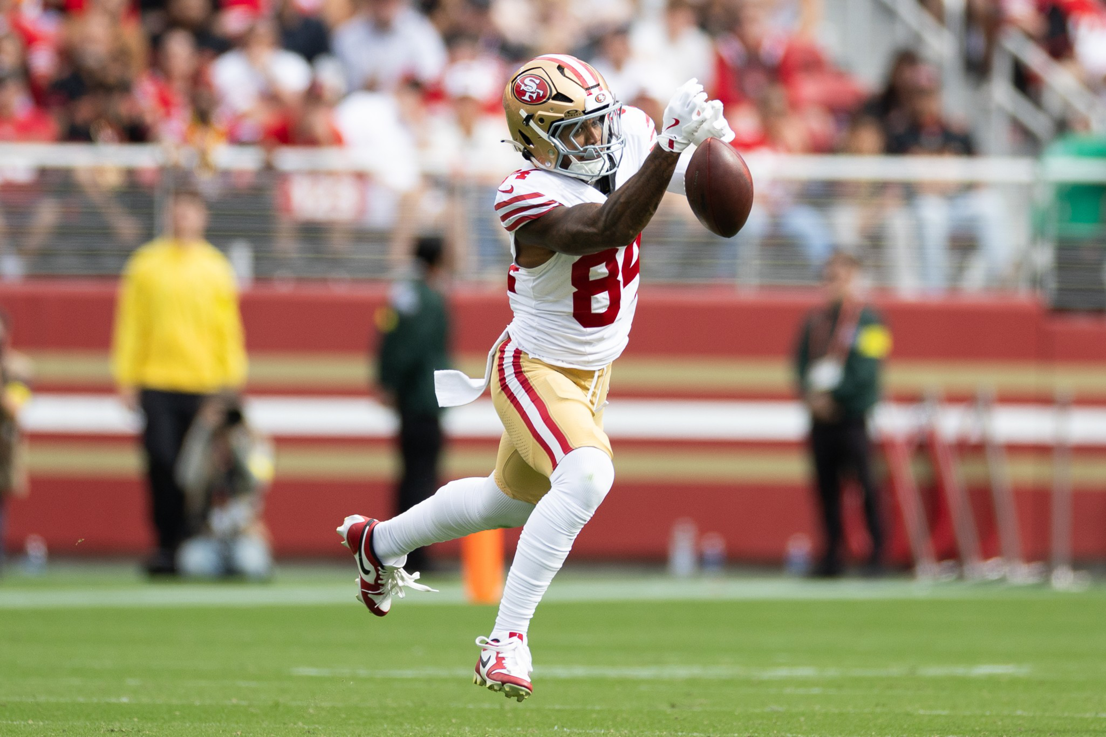 A football player in white and gold uniform leaps to catch a football midair on a green field with blurred spectators in the background.