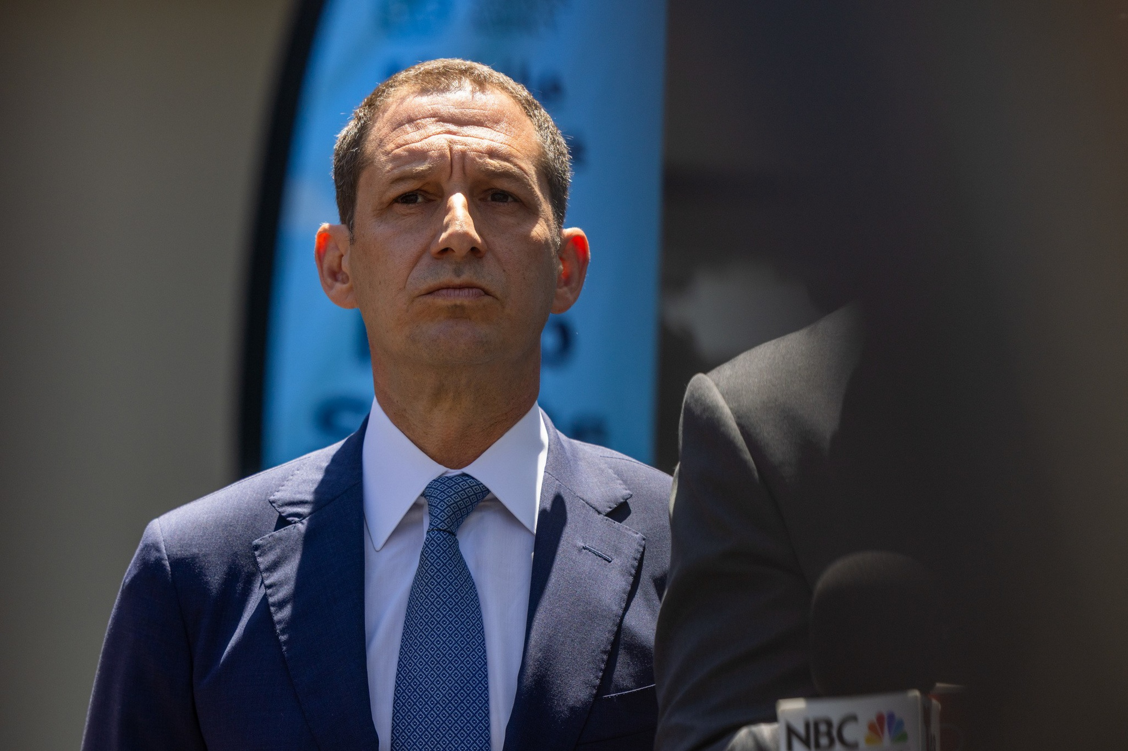 A man in a blue suit and patterned blue tie stands outdoors, looking serious, with part of another person and an NBC microphone visible nearby.
