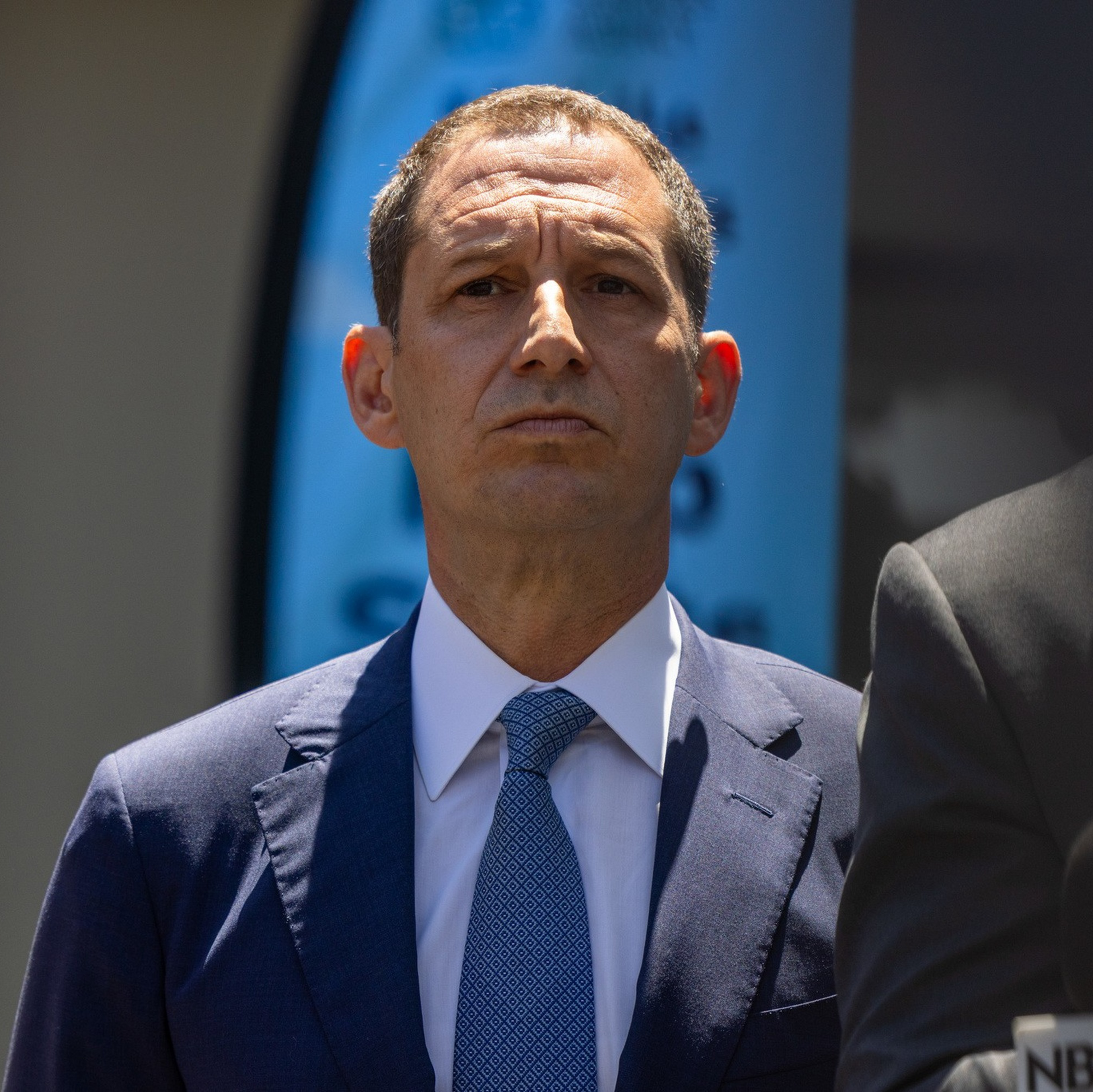 A man in a blue suit and patterned blue tie stands outdoors, looking serious, with part of another person and an NBC microphone visible nearby.