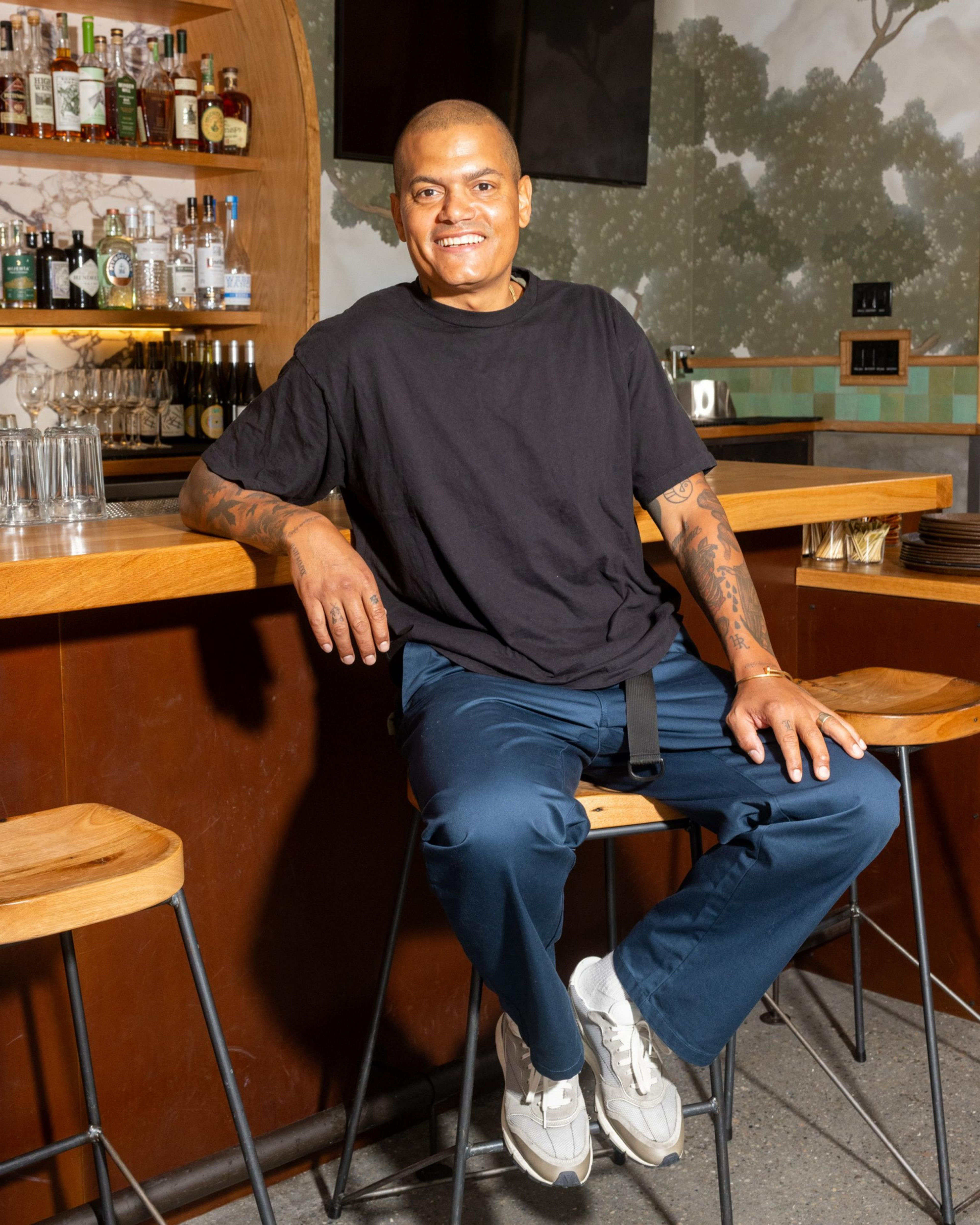 A person with a shaved head and tattoos is sitting on a bar stool, smiling. They're wearing a black t-shirt, blue pants, and white sneakers, in front of a well-stocked bar.