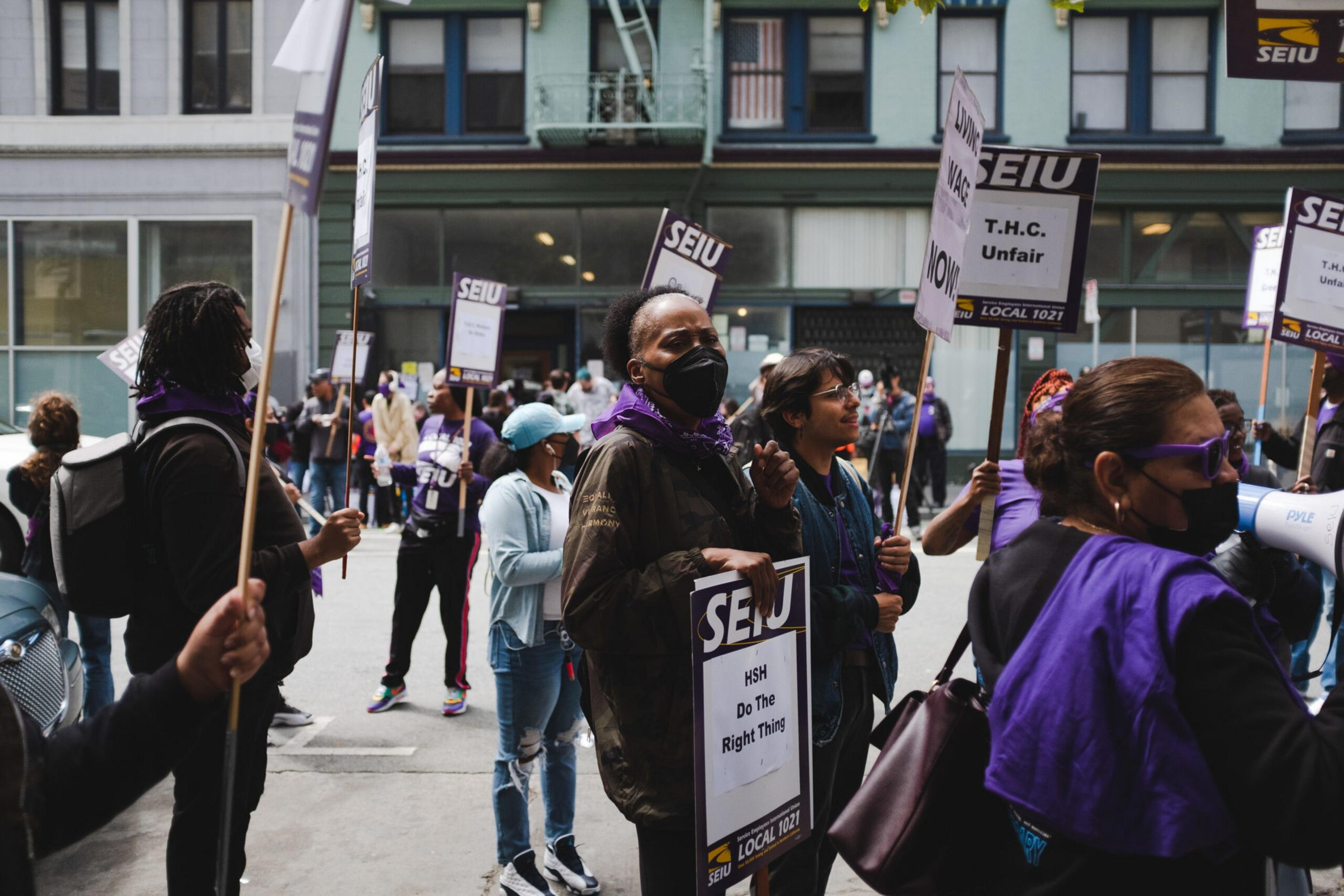 A group of masked protesters holds signs with messages like "SEIU," "HSH Do The Right Thing," and "T.H.C. Unfair" during a street demonstration.