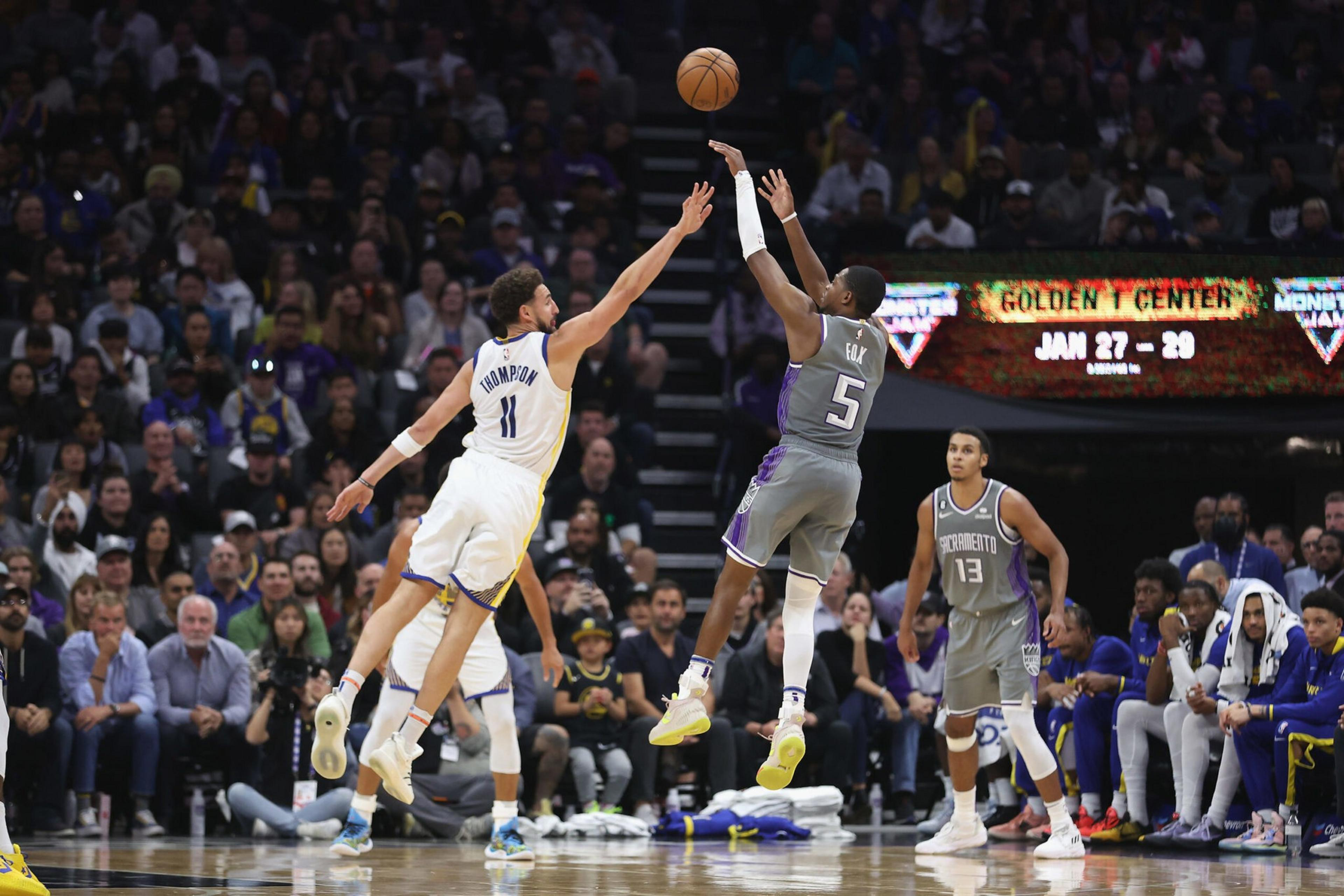 Two basketball players jump as one shoots the ball while the other tries to block in front of an audience and nearby teammates.