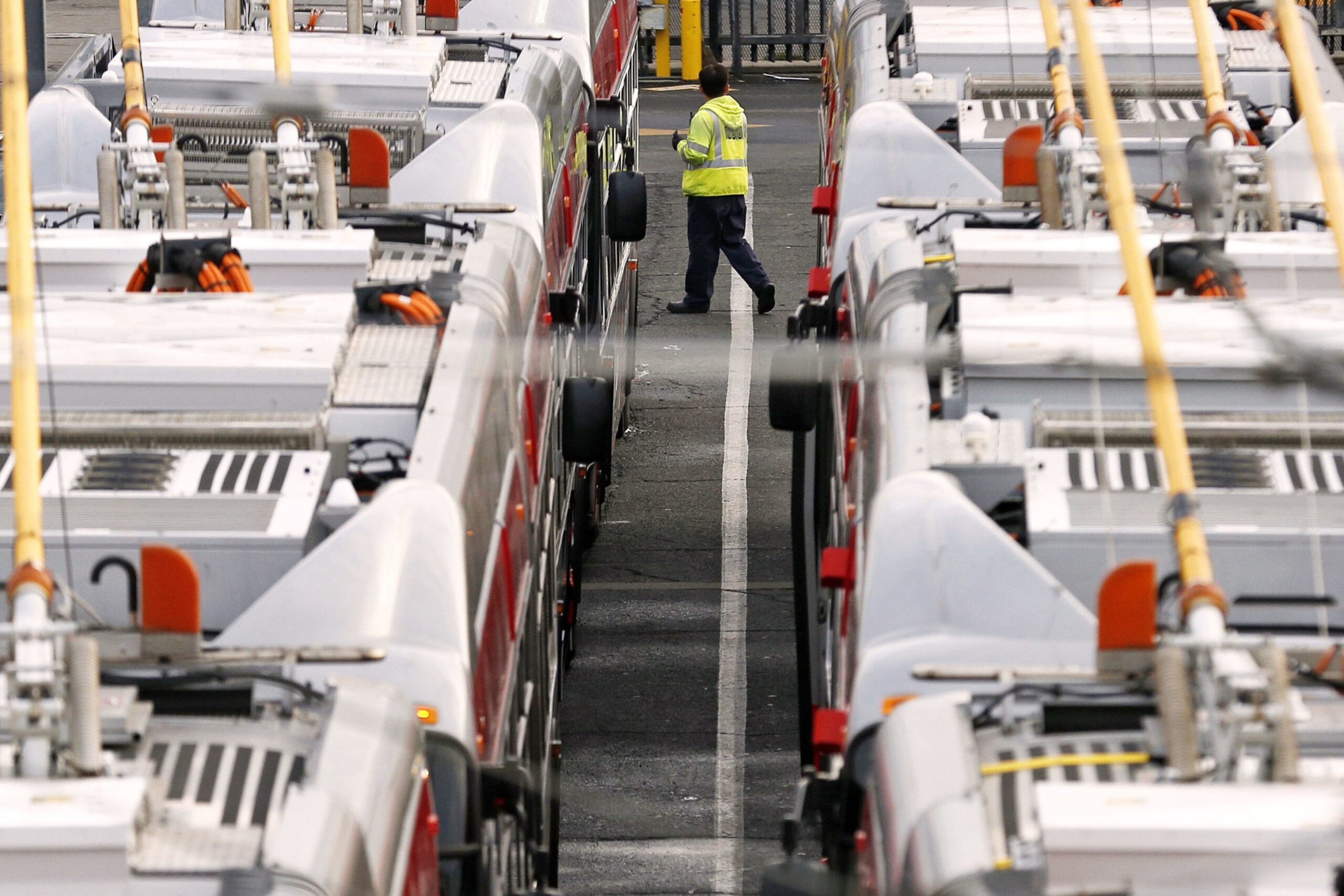 A person in a yellow safety vest walks between two rows of parked gray and red buses at a transit depot.