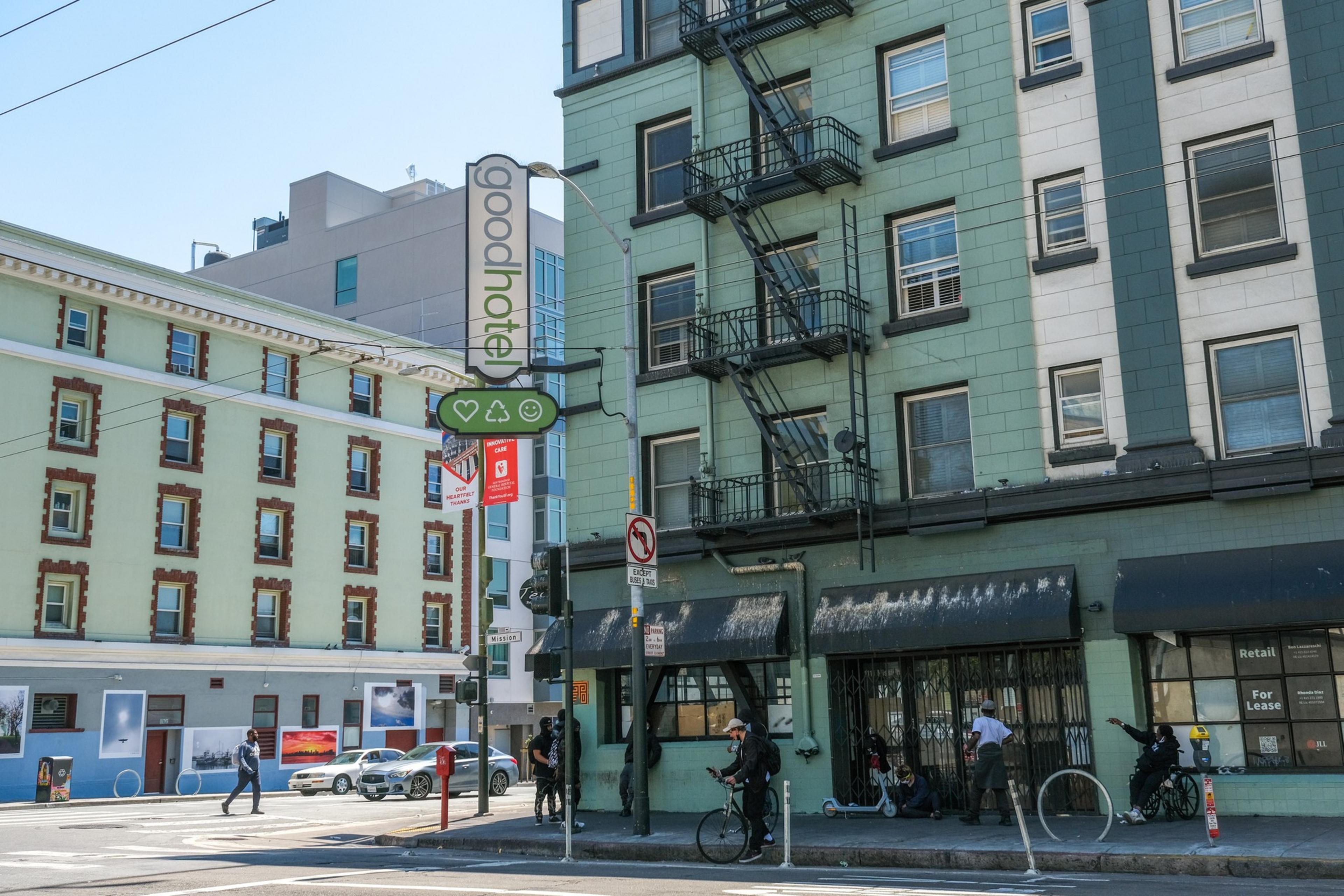 A street corner with a green building featuring a fire escape, several people on bikes and scooters, a "goodpotato" sign, and a "Retail For Lease" window.