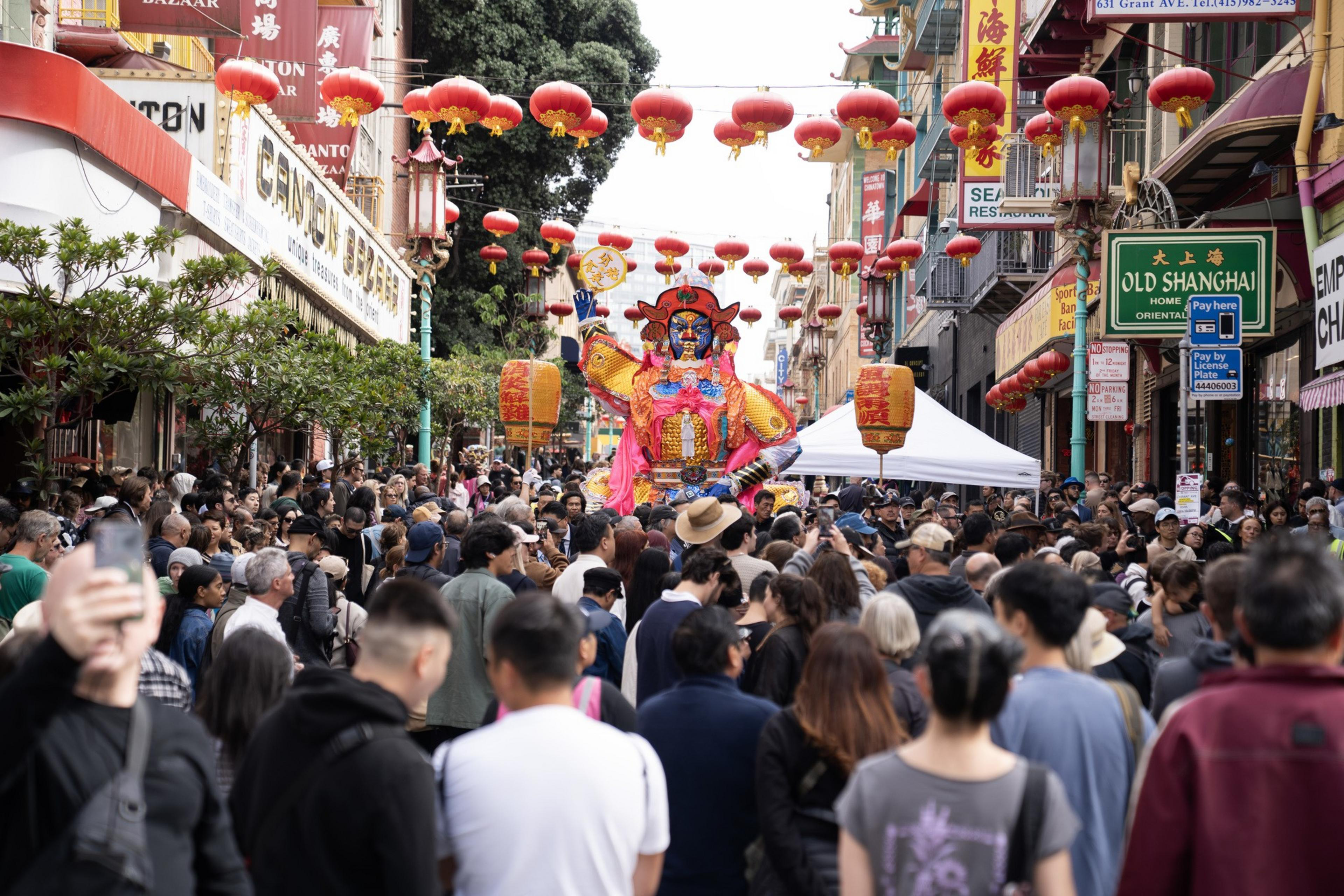A crowded street decorated with red Chinese lanterns features a colorful, large traditional figure during a festive celebration.