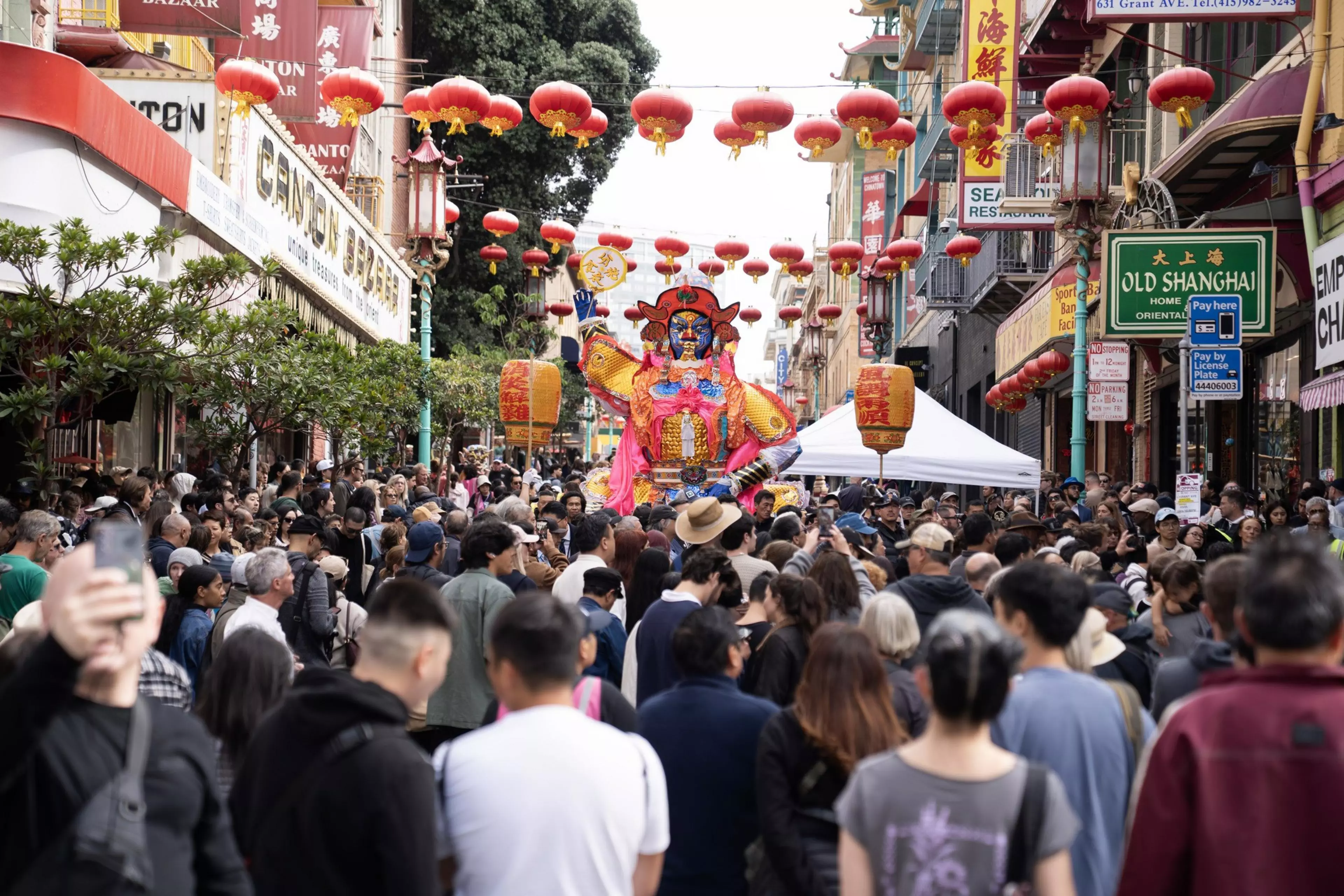 A crowded street decorated with red Chinese lanterns features a colorful, large traditional figure during a festive celebration.