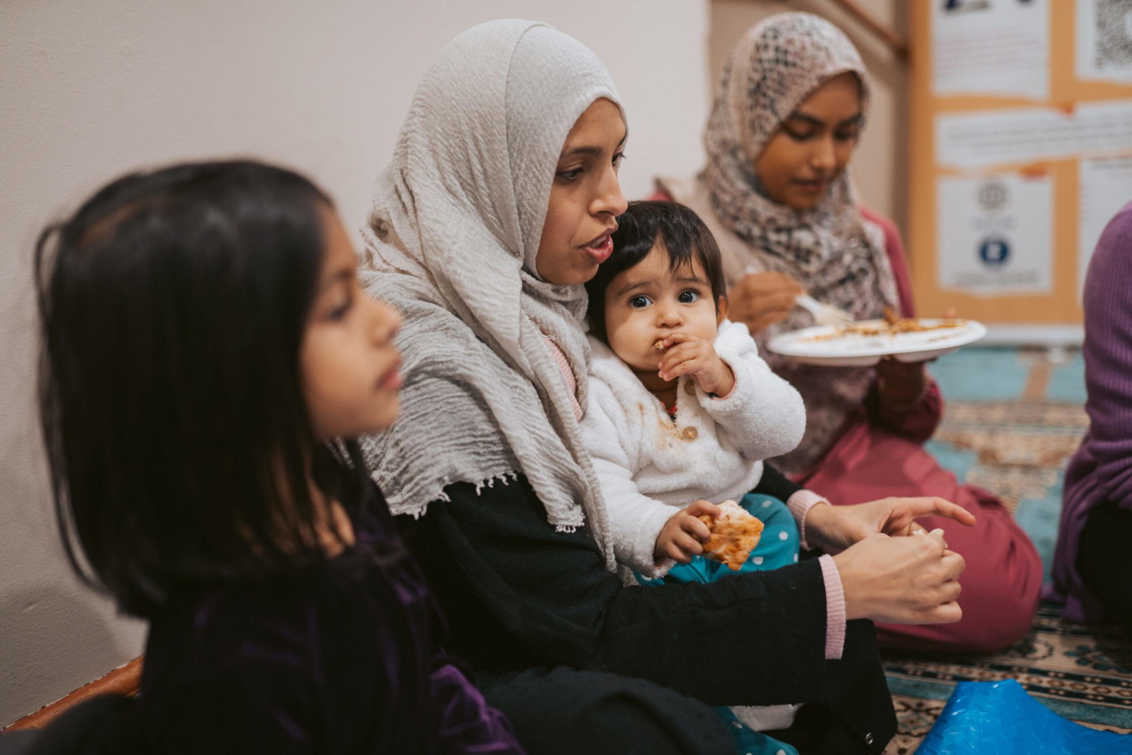 A woman in a hijab holds a baby eating food, while two other girls sit nearby on a patterned carpet with plates of food.