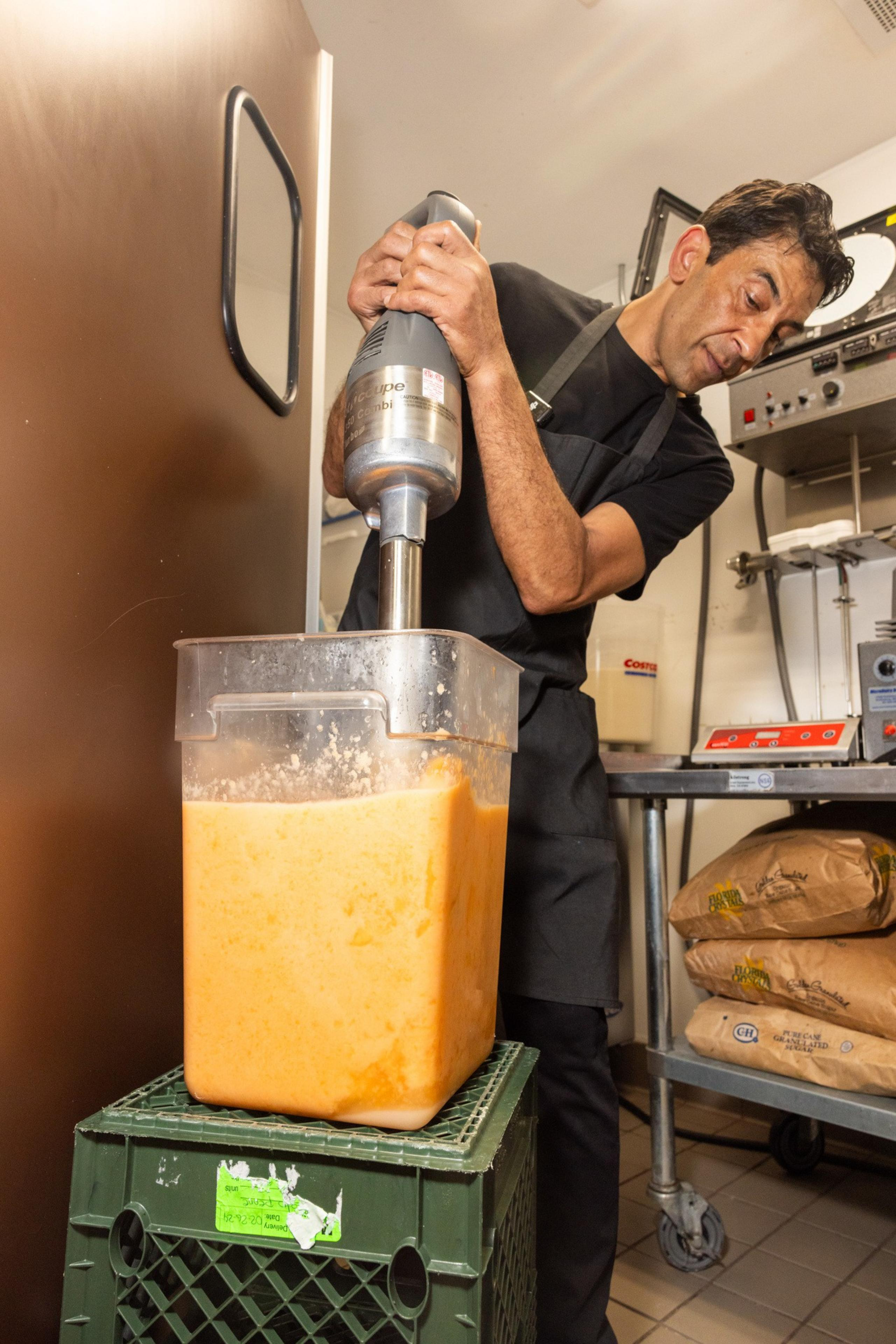 A person in a black apron and shirt uses an immersion blender to mix a large container of orange liquid. The container is placed on a green crate in a kitchen.