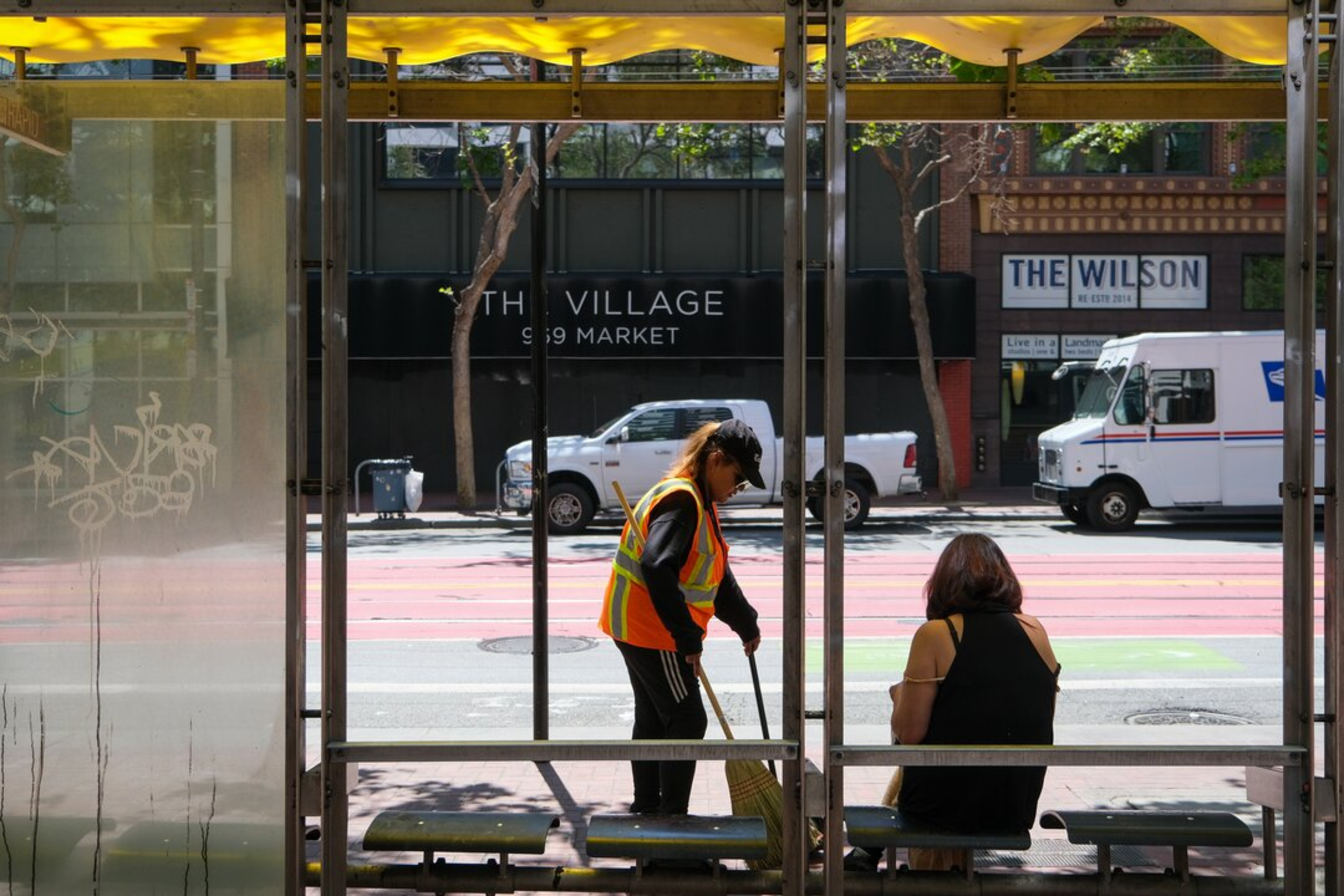 A worker in a safety vest sweeps the floor of a bus stop while a woman sits on the bench facing the street with parked vehicles and storefronts behind.