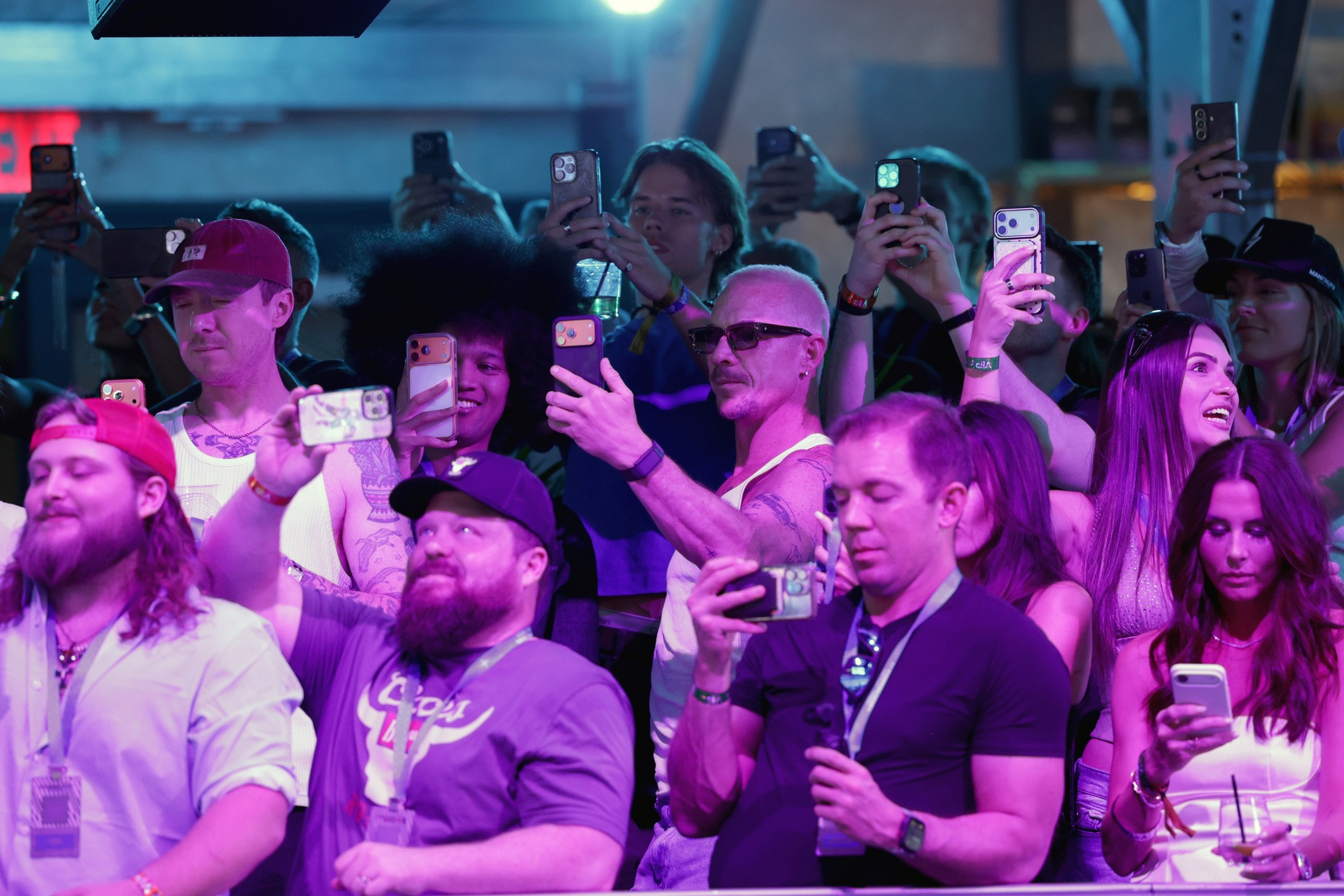 A diverse group of people at an event take photos or videos with their smartphones under purple and blue lighting.