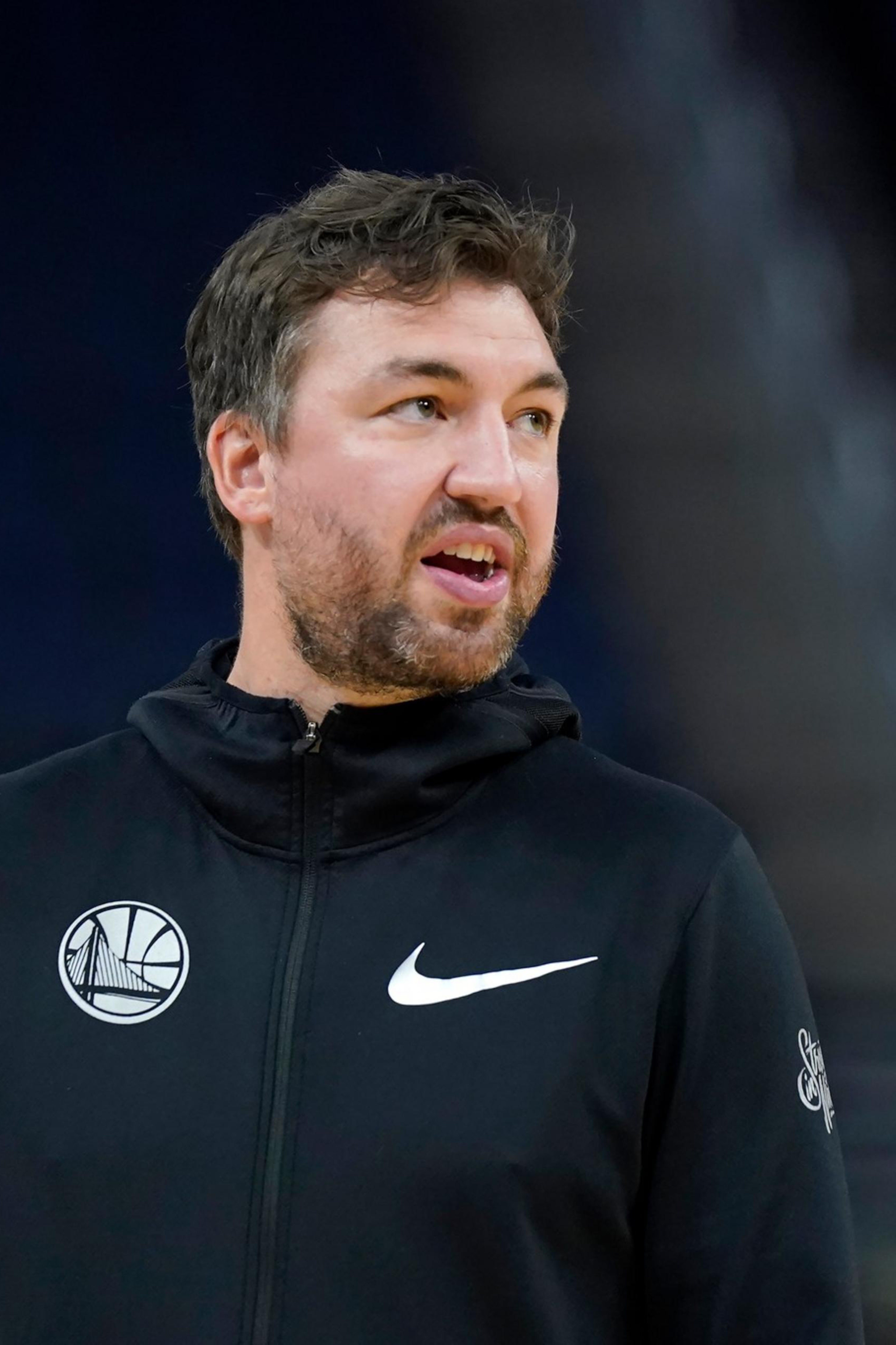 A man with short dark hair and a beard wears a black Nike hoodie with a Golden State Warriors logo, standing in a dimly lit indoor area.