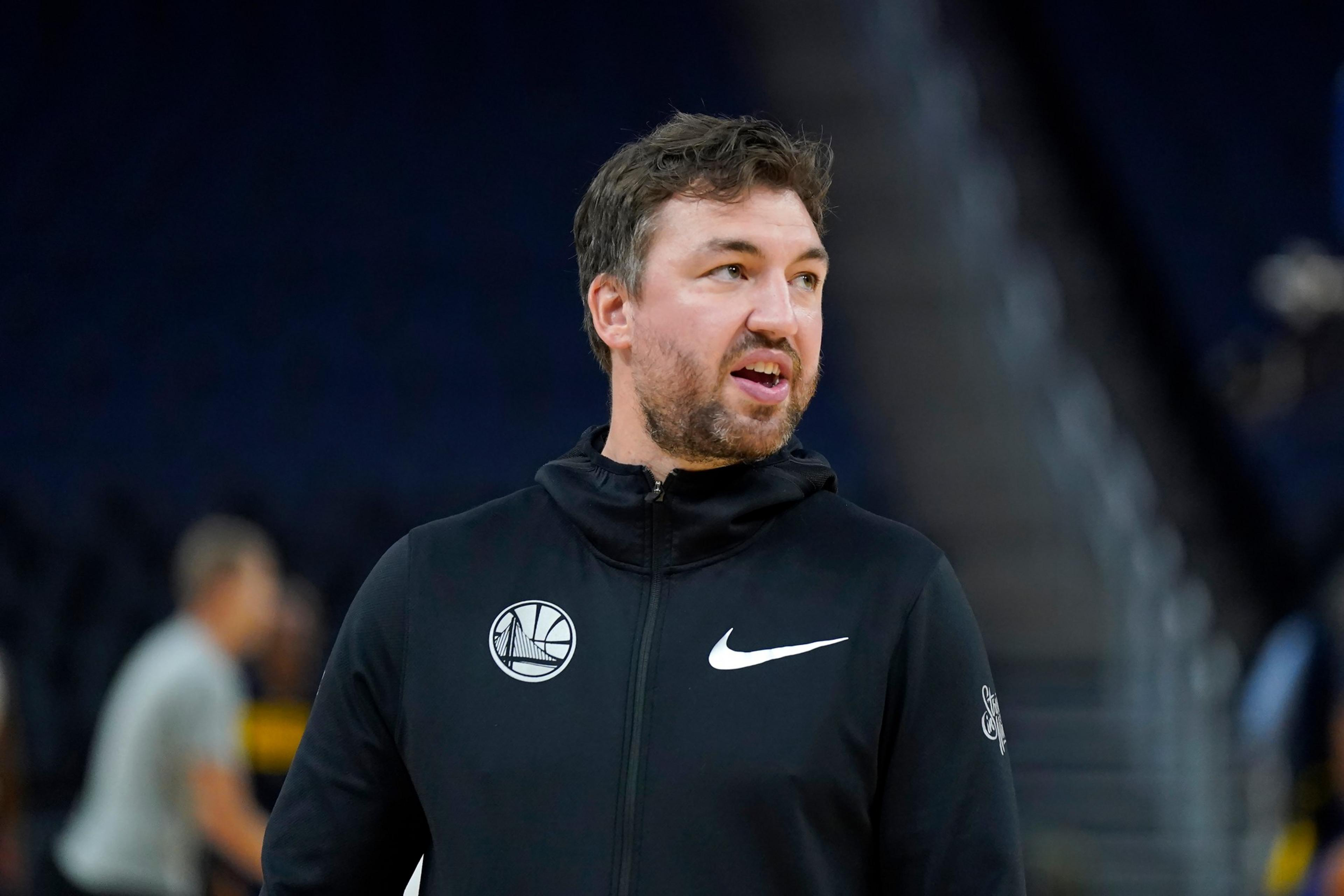 A man with short dark hair and a beard wears a black Nike hoodie with a Golden State Warriors logo, standing in a dimly lit indoor area.