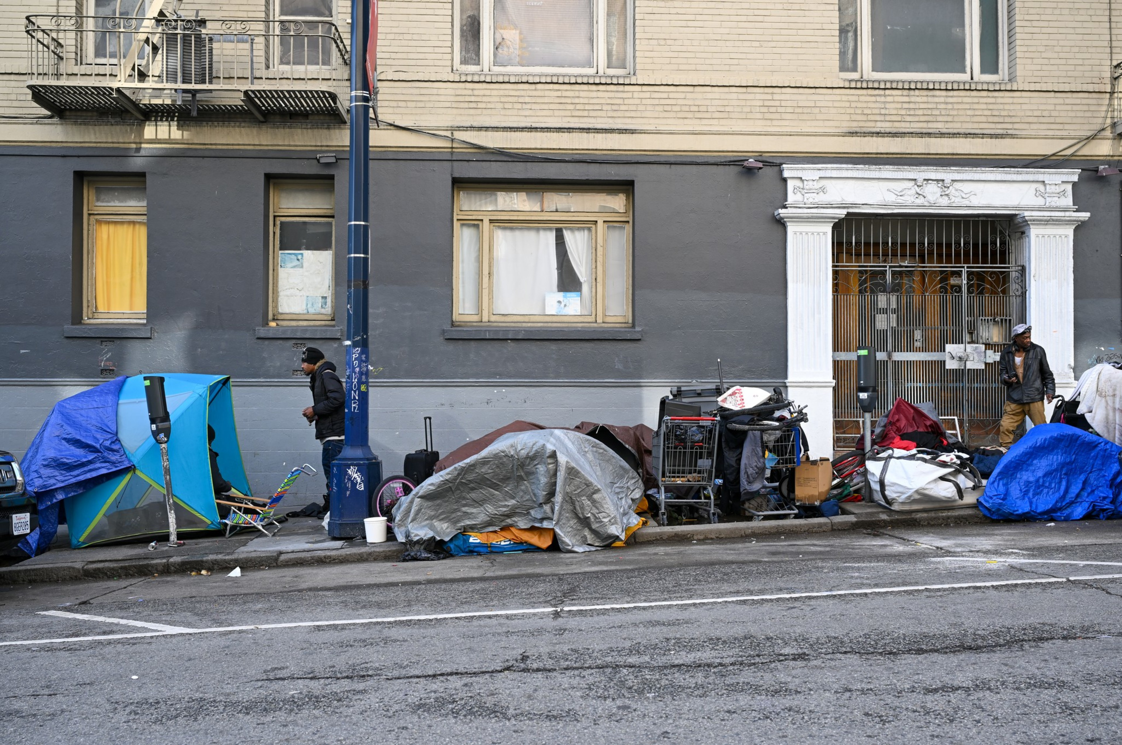 Several tents and tarps cover belongings on the sidewalk in front of a building, with two people standing near the makeshift shelters.