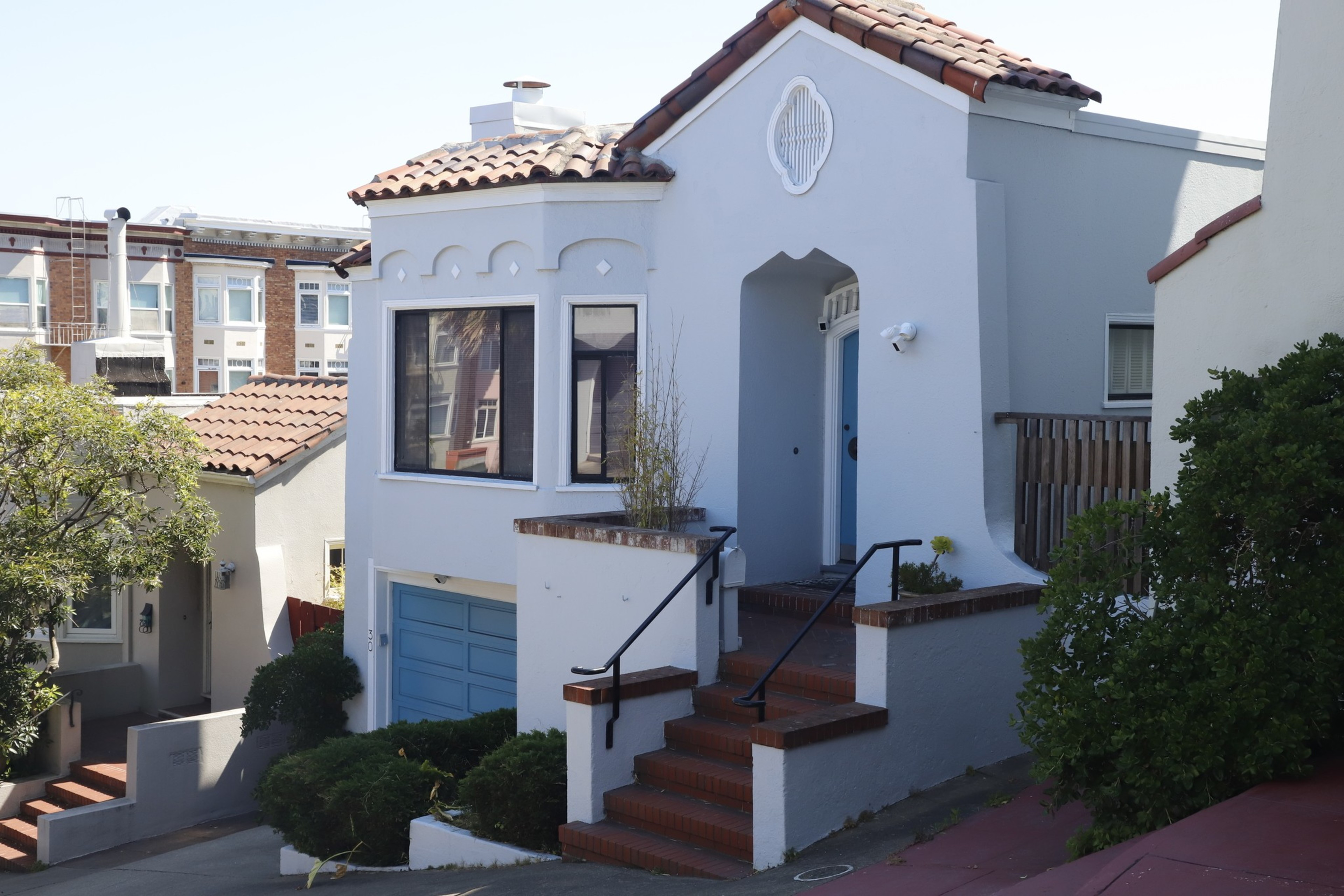The image shows a small two-story white house with a terracotta tiled roof, large front window, blue garage door, and steps leading to a blue front door. Surrounding greenery includes a small tree and bushes.