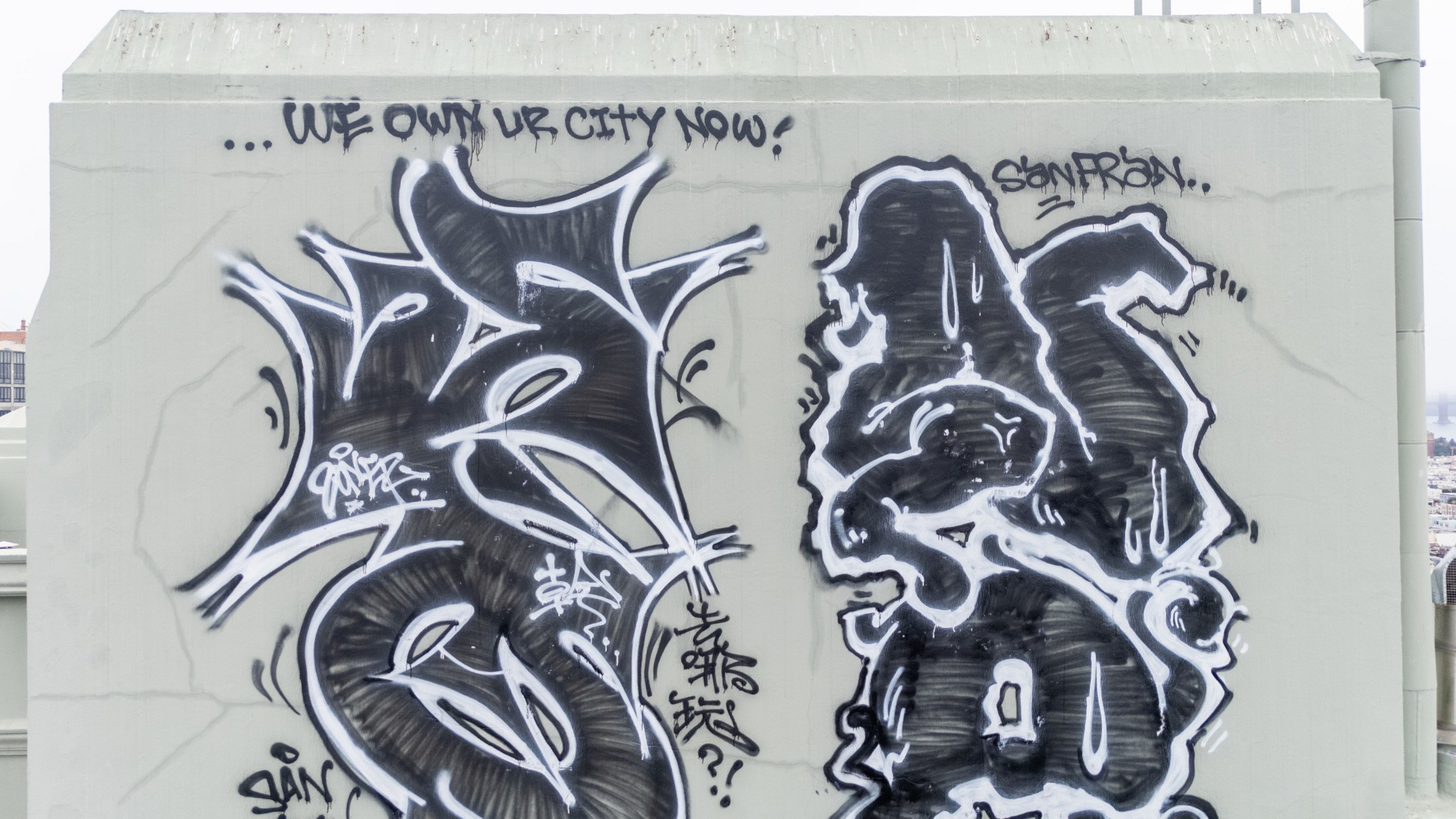 Black-and-white graffiti on a concrete wall with abstract shapes and the text “WE OWN UR CITY NOW!” written above.