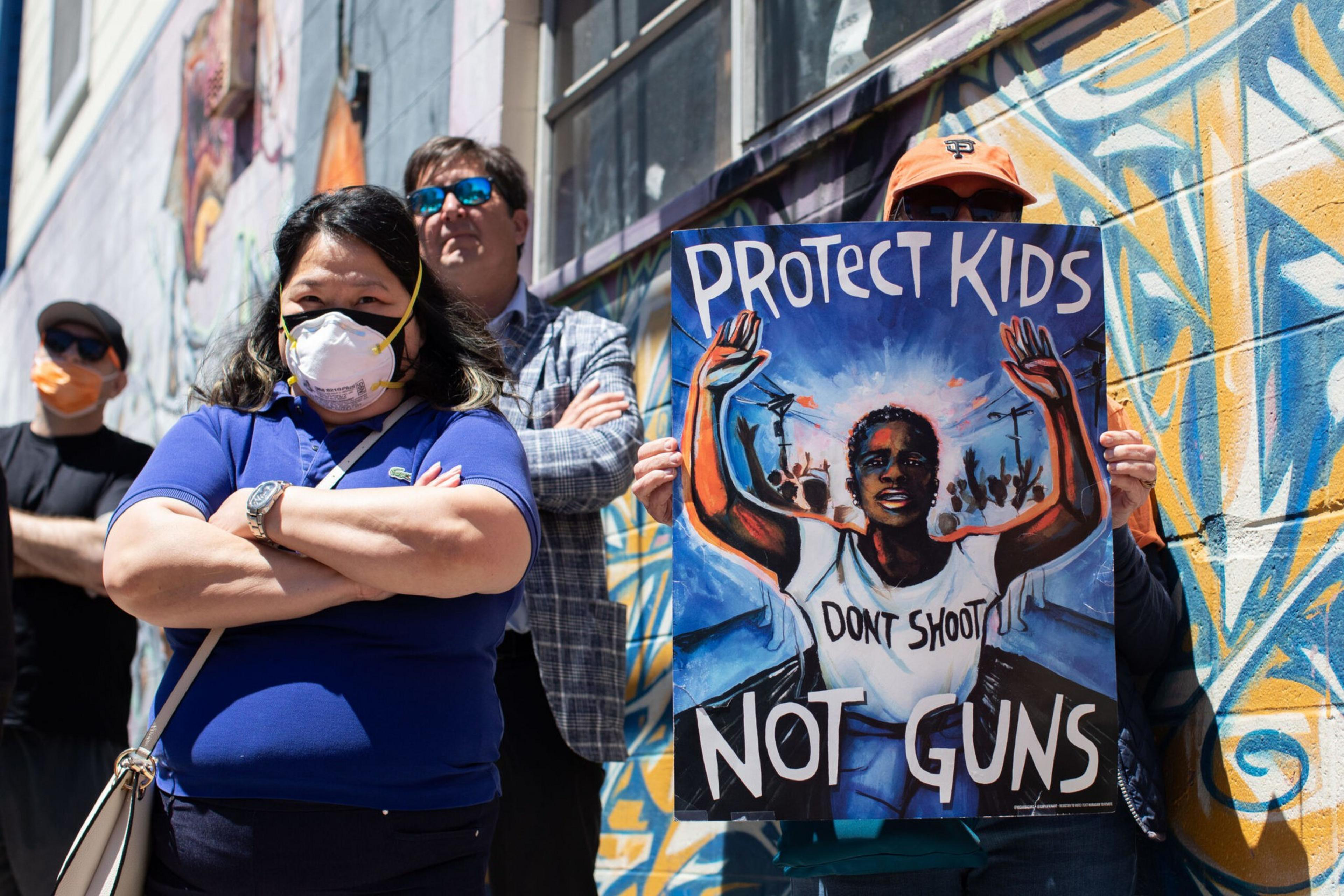 A group of people stand by a graffiti wall, one holding a colorful sign that reads "Protect Kids, Don't Shoot, Not Guns." Most wear masks.