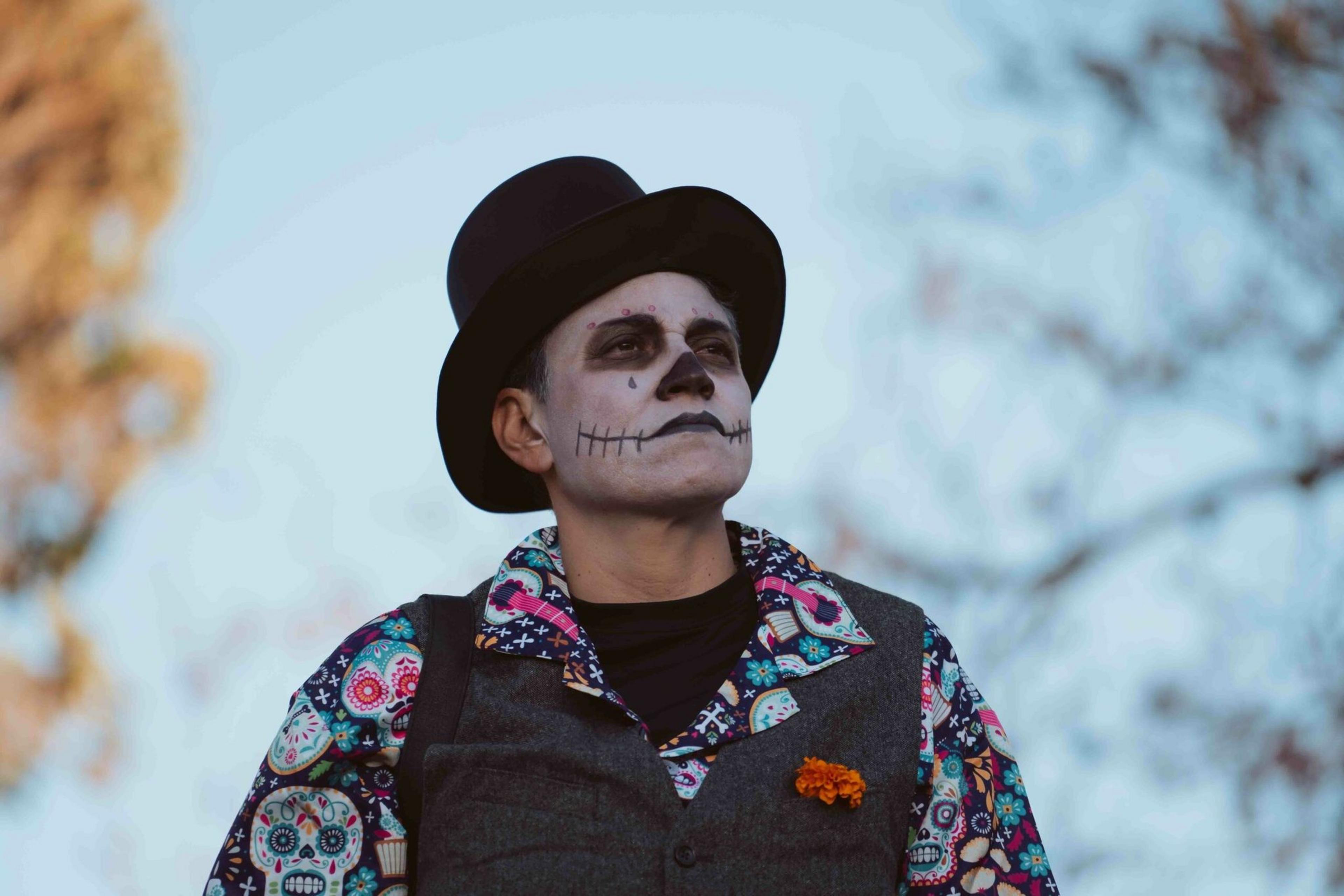 A person wears skeleton face paint, a black top hat, a colorful skull-patterned shirt, and a gray vest with an orange flower pinned to it.