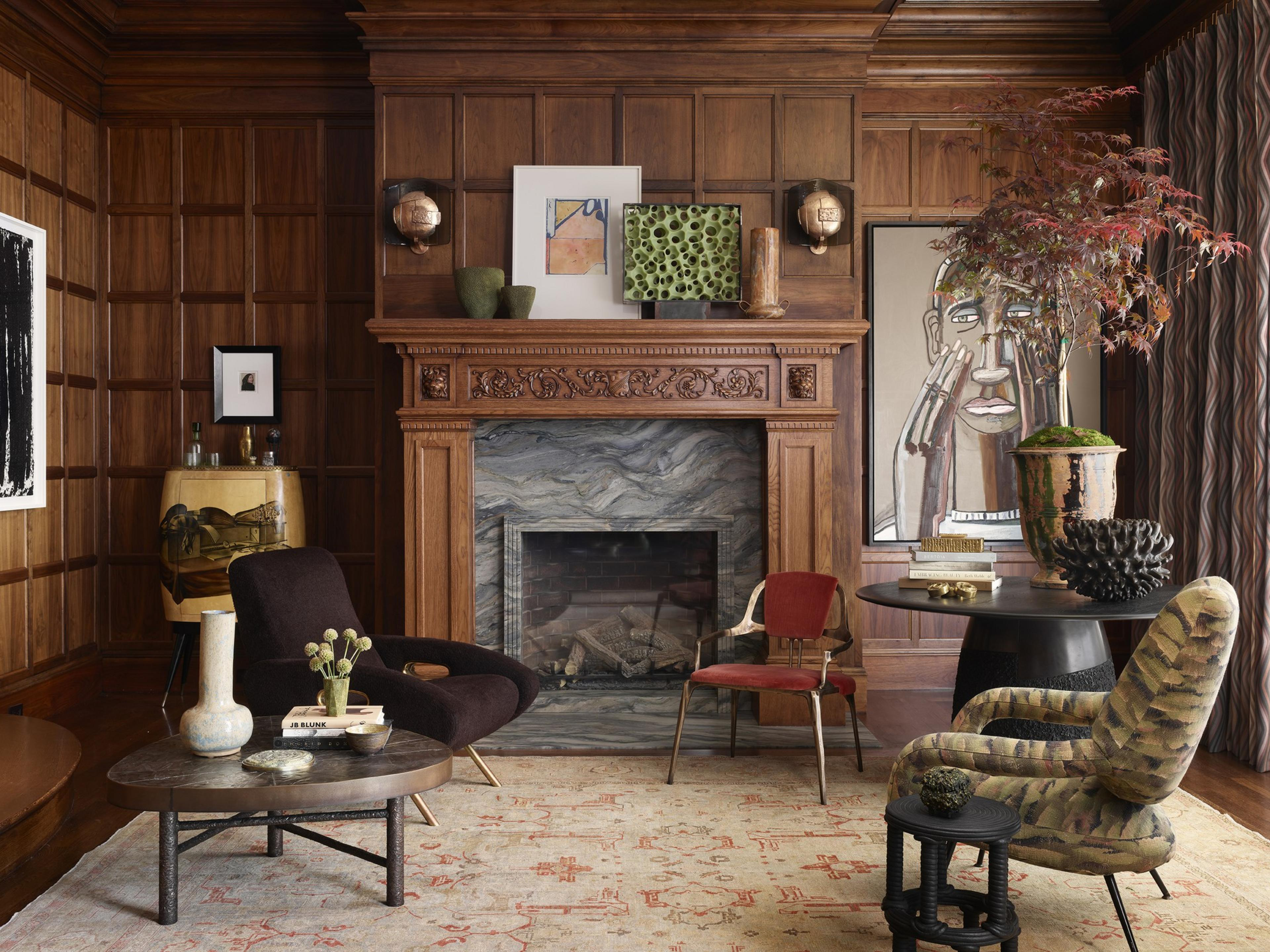 A wood-paneled room with a carved fireplace, various art pieces, a dark chair, a red chair, patterned upholstery, a round table, and a light area rug.