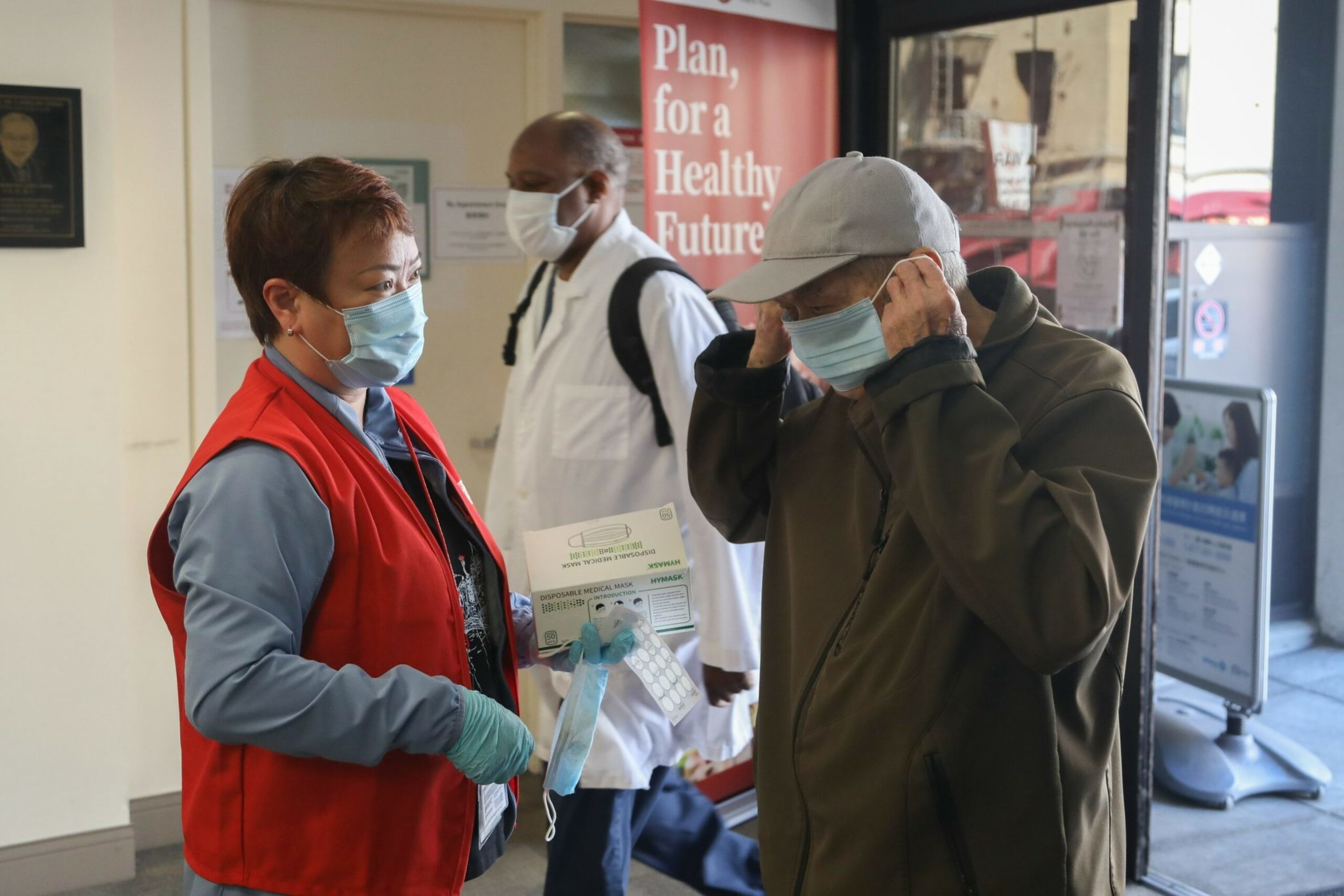 A woman in a red vest hands masks to an elderly man adjusting his mask, while a masked man in a white coat walks past a "Plan for a Healthy Future" sign.