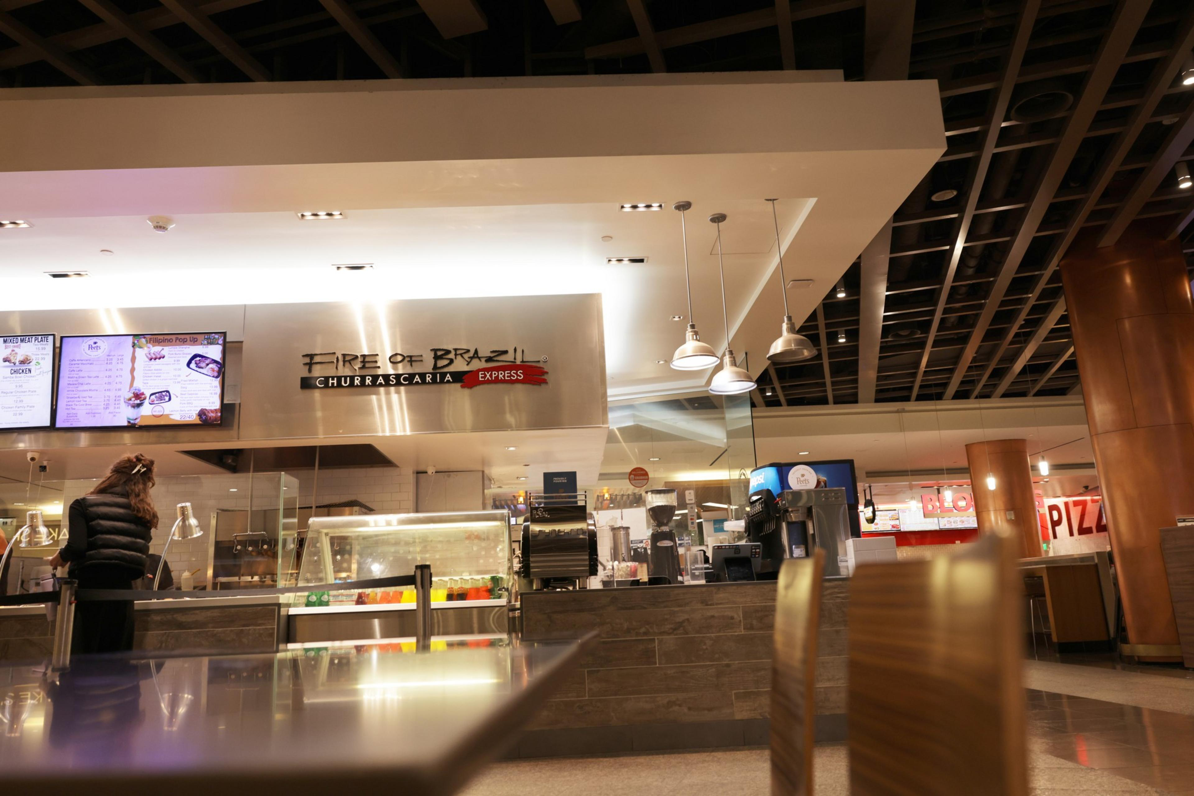An indoor food court area featuring a Brazilian restaurant, with a customer at the counter and modern lighting overhead.