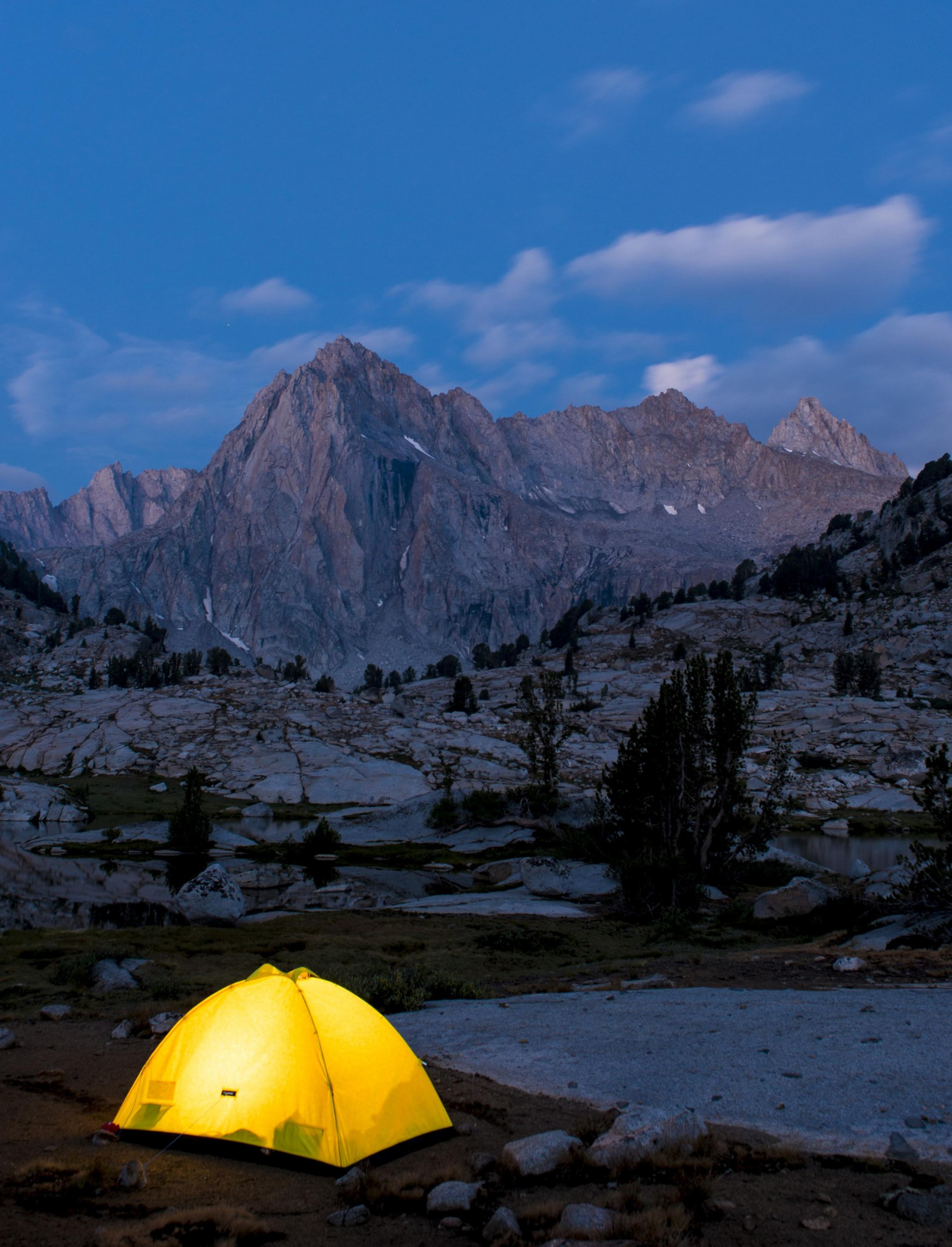 A glowing yellow tent sits on rocky terrain under a clear dusk sky, with majestic mountains and some scattered clouds in the background.
