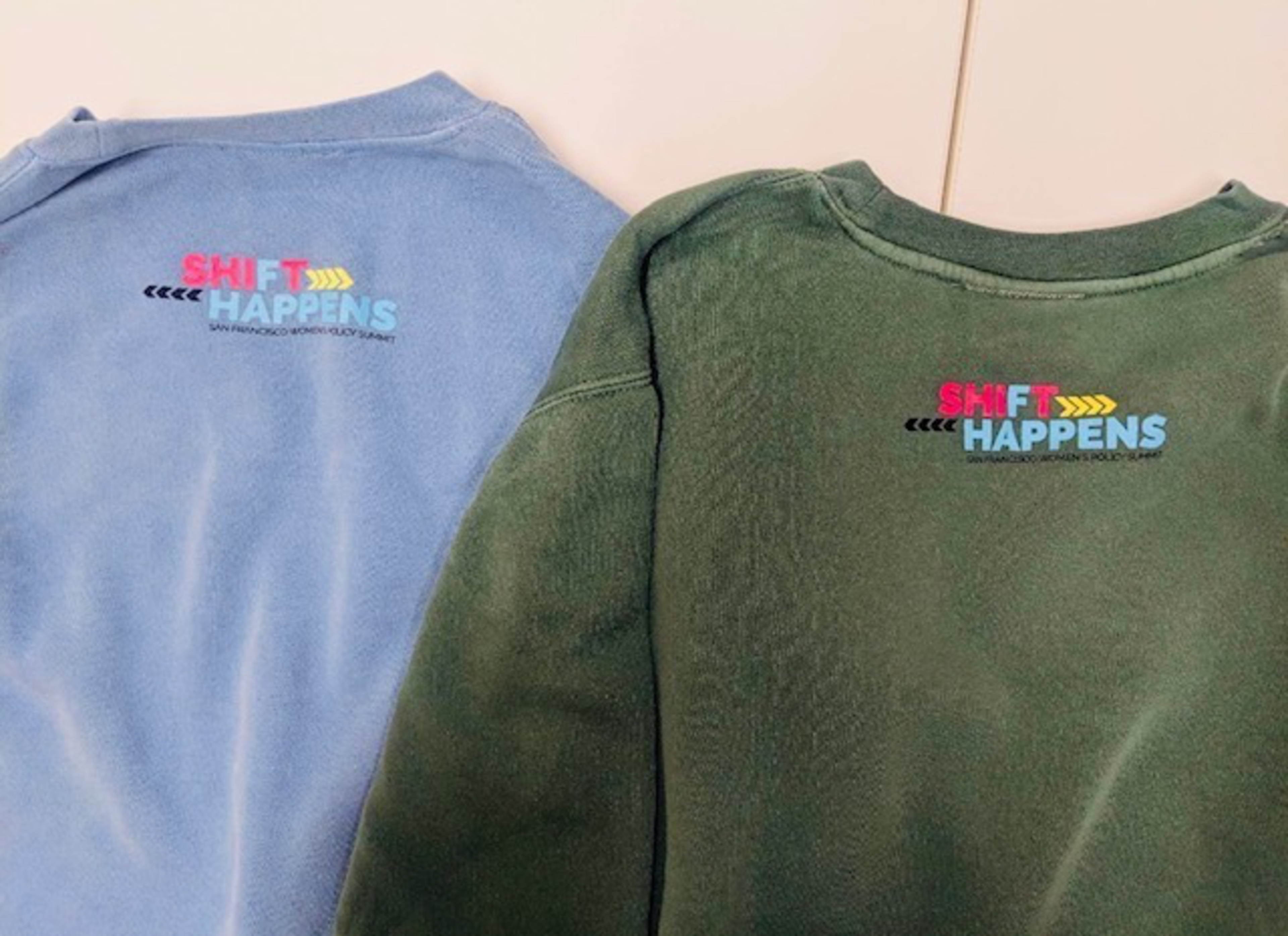 Two sweatshirts, one blue and one green, both have the colorful text "SHIFT HAPPENS" and "SAN FRANCISCO WOMEN'S POLICY SUMMIT" on their backs near the neckline.