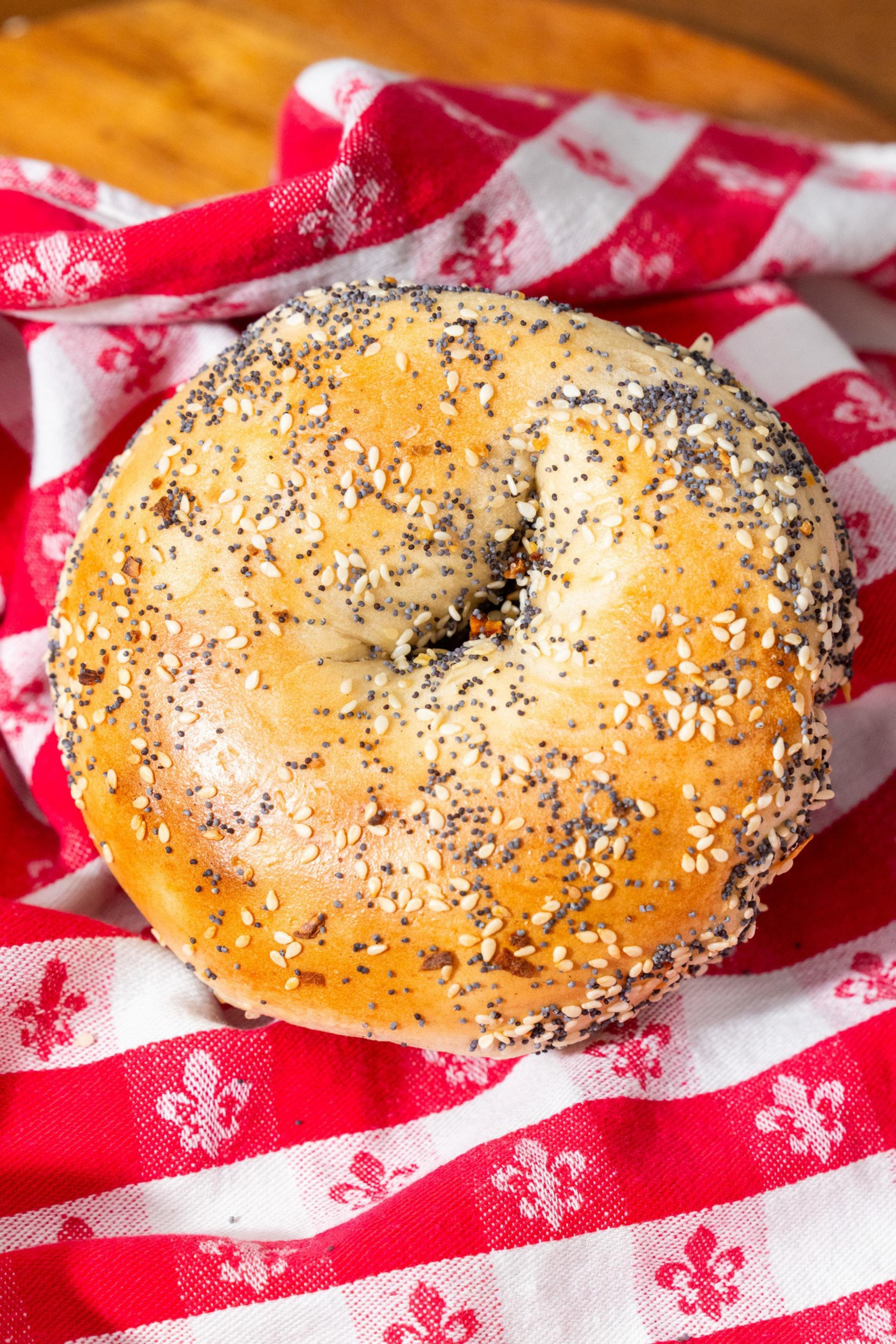 A golden bagel topped with sesame seeds, poppy seeds, and seasoning rests on a red and white checkered cloth with fleur-de-lis patterns.