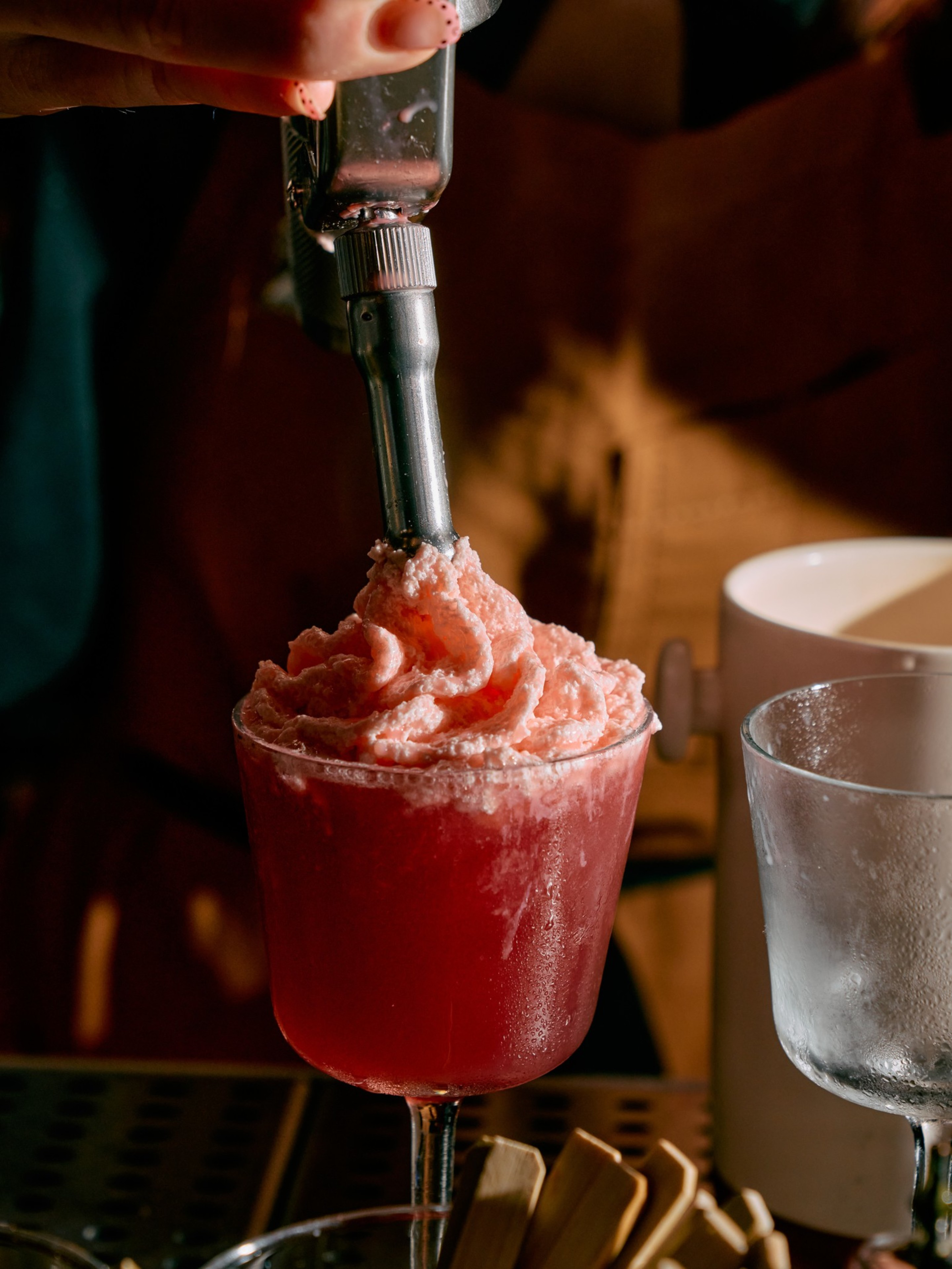 A hand uses a metal scoop to add a pink, fluffy, icy mixture on top of a red drink in a stemmed glass, with a frosted glass nearby.