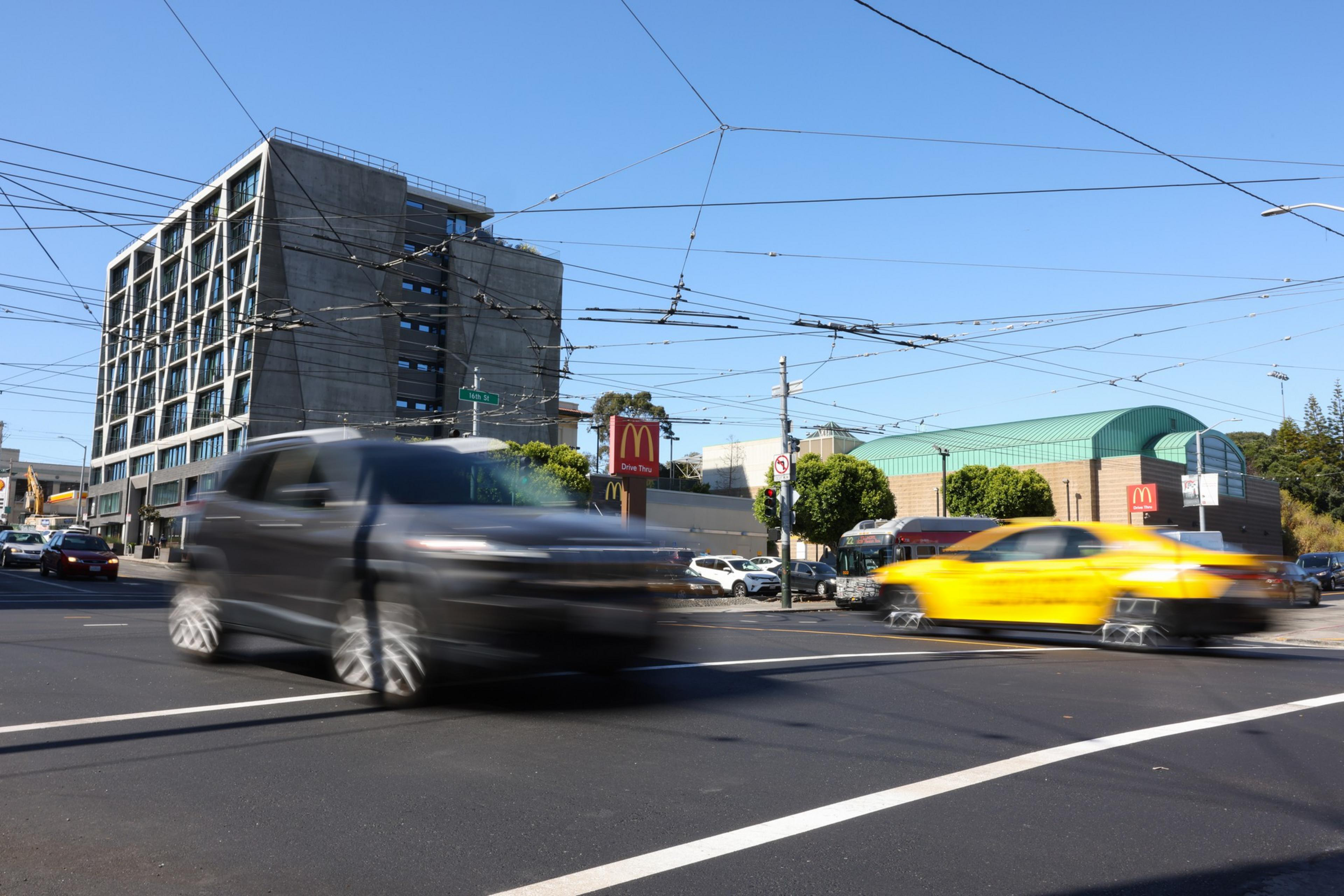A busy urban intersection has blurred vehicles speeding by, with a McDonald's restaurant and a large building with a grid-like facade in the background.
