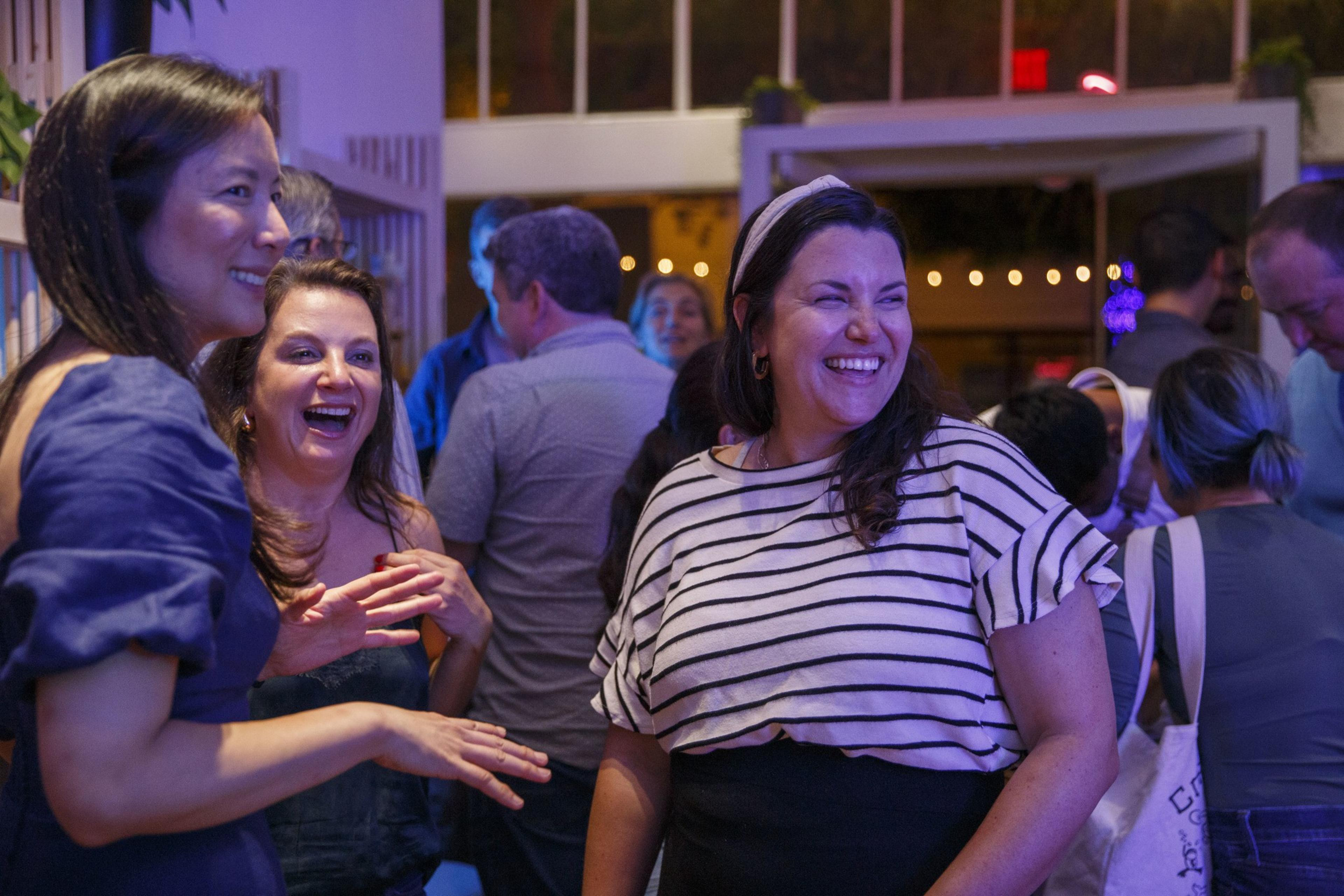 Three women standing in a bar while close together look into the distance and have a laugh.