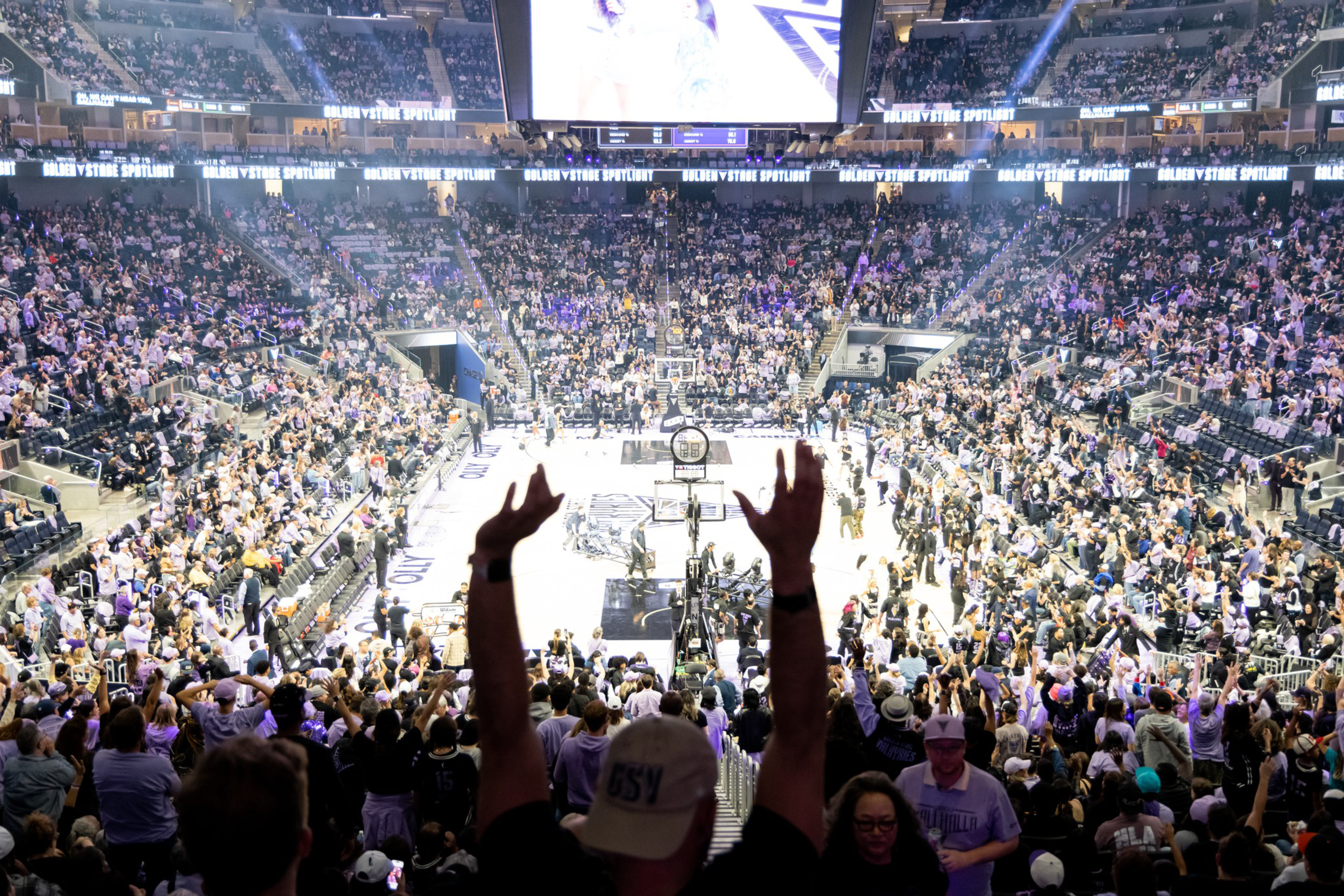 A large crowd of fans in an arena cheer as a basketball game takes place on the court below, with bright lights and a scoreboard overhead.