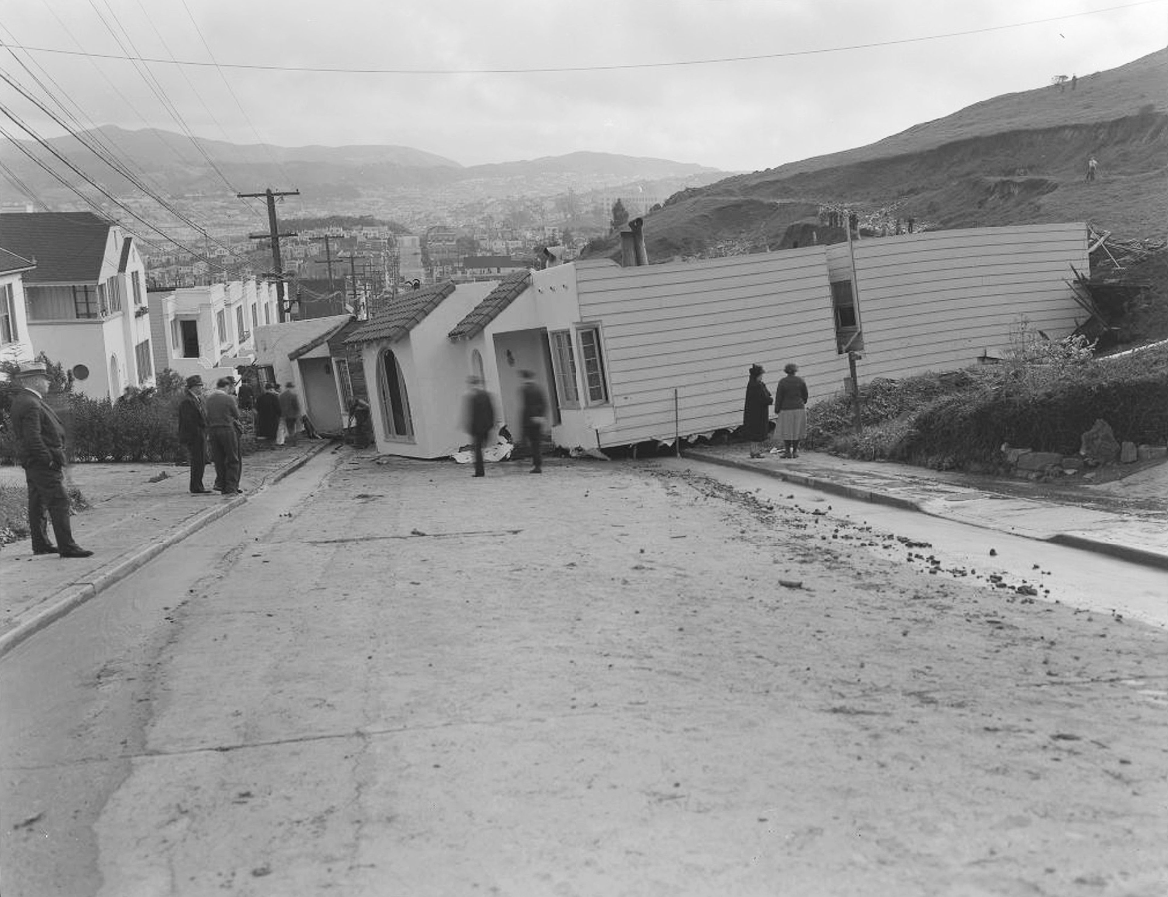 A tilted house blocks a street after being displaced, with several people standing nearby observing the damage and the surrounding houses intact.