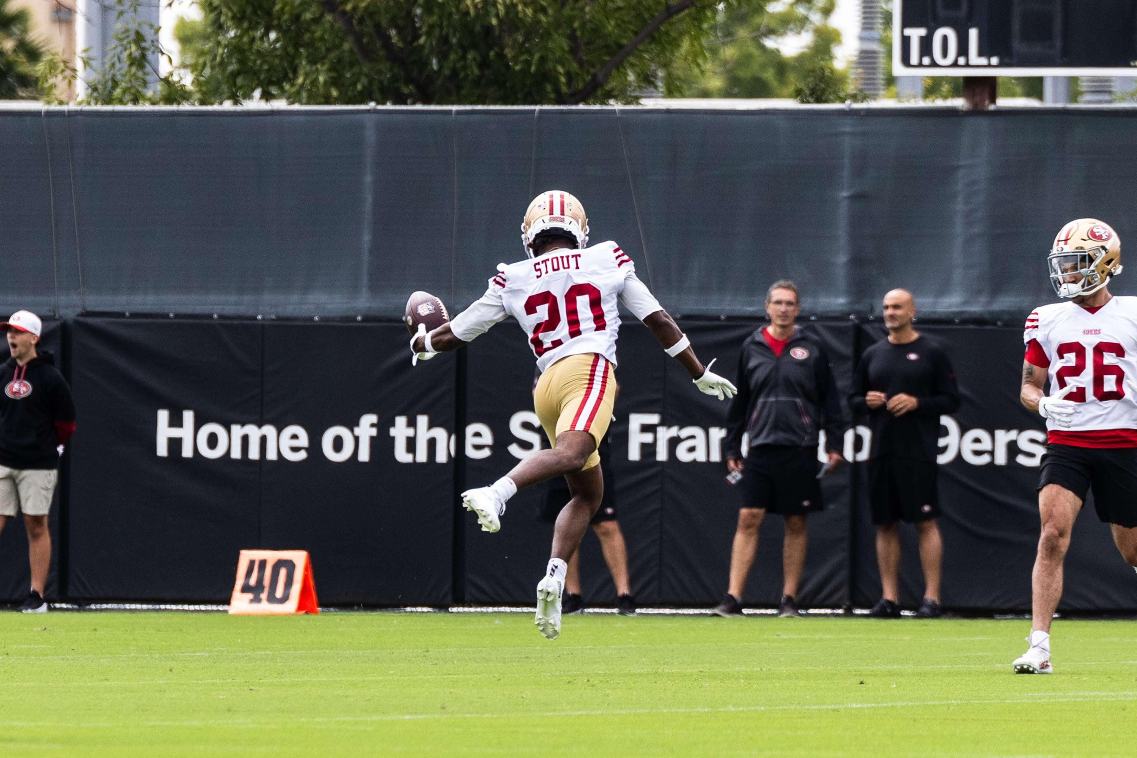 A football player in a white and red uniform jumps with a ball in hand on a field. A sign reads &quot;Home of the San Francisco 49ers&quot; in the background.