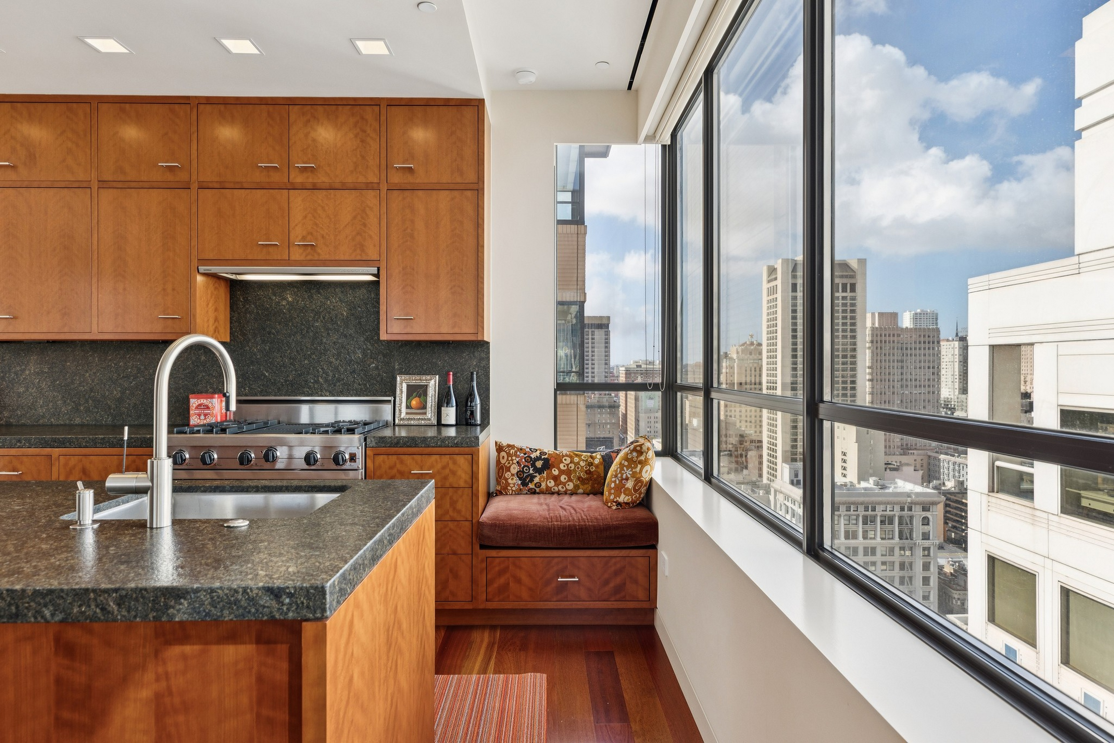 A modern kitchen with wooden cabinets, a black stone countertop island, a stainless steel stove, a window bench with cushions, and large city-view windows.