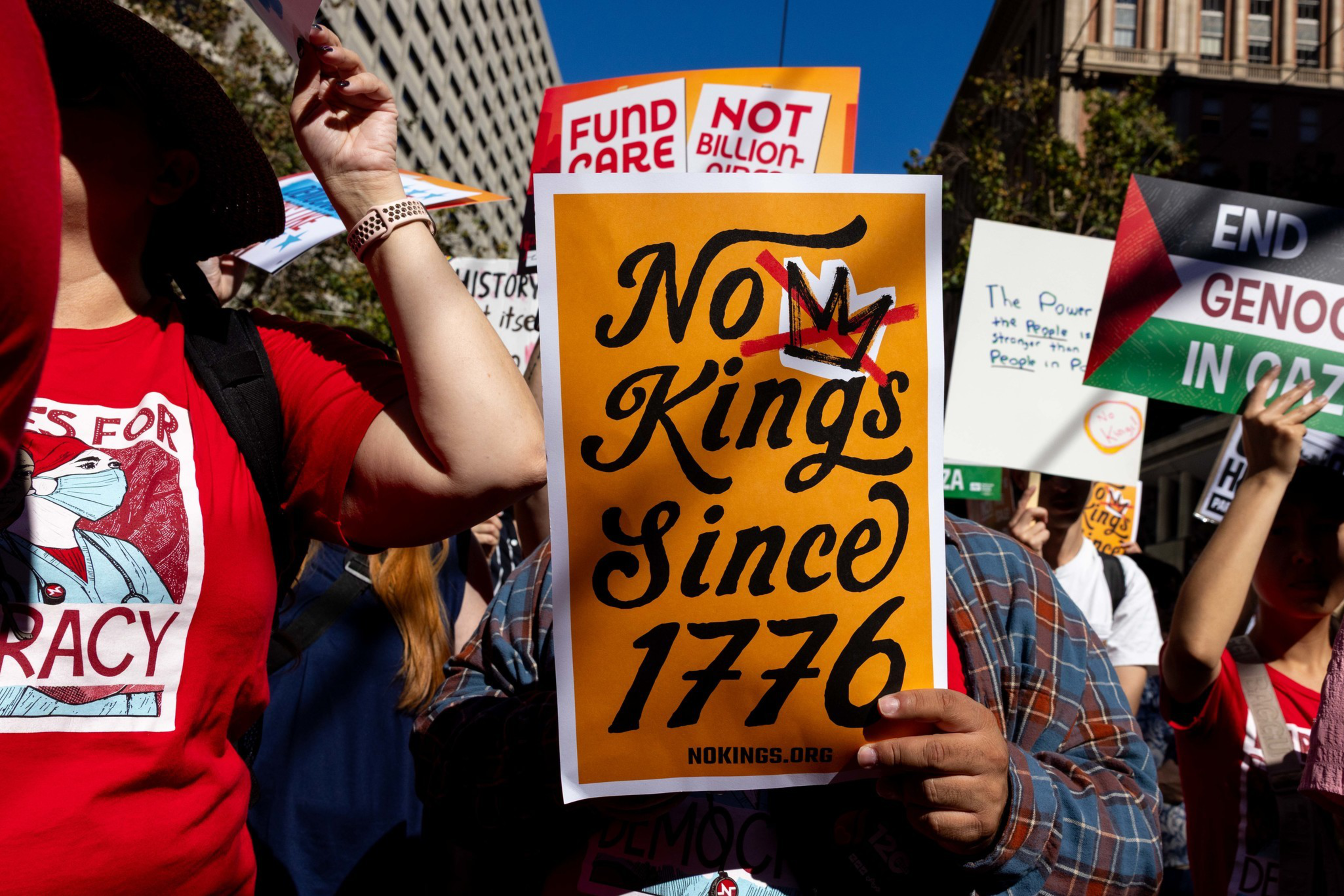 A person holds a sign reading “No Kings Since 1776" with a crown illustration, surrounded by others holding protest signs about democracy and Gaza.