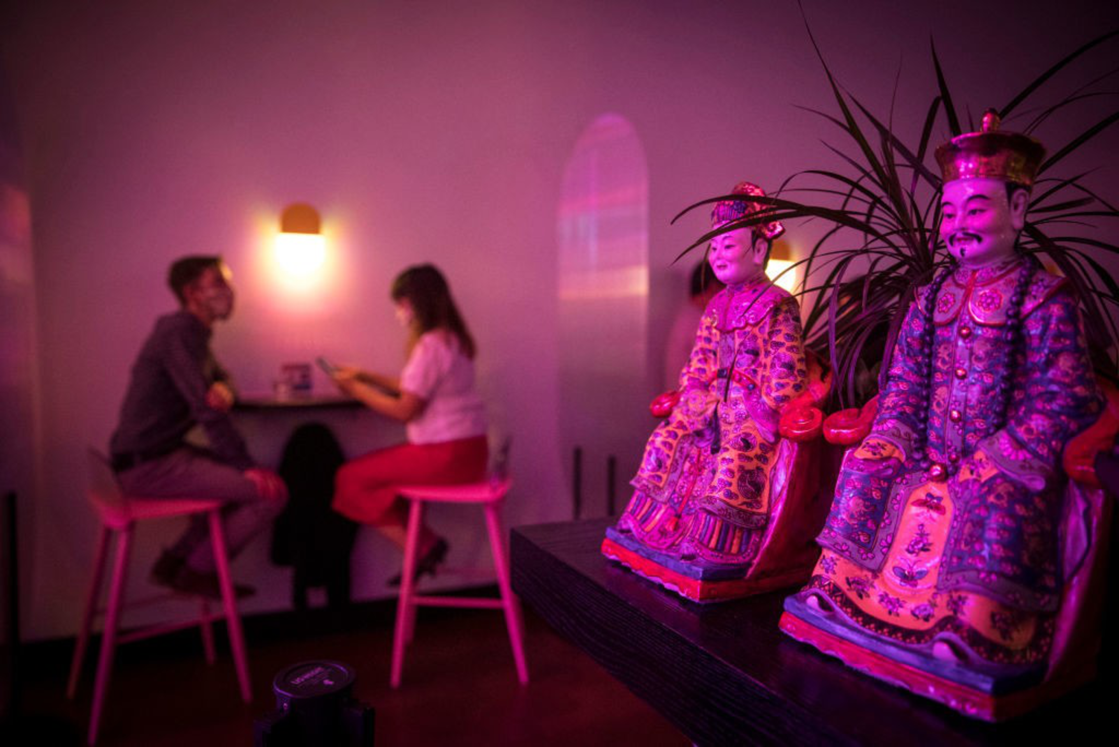 Two colorful, detailed figurines of traditionally dressed figures sit on a shelf, glowing under purple-pink lighting, while two people chat at a table in the background.