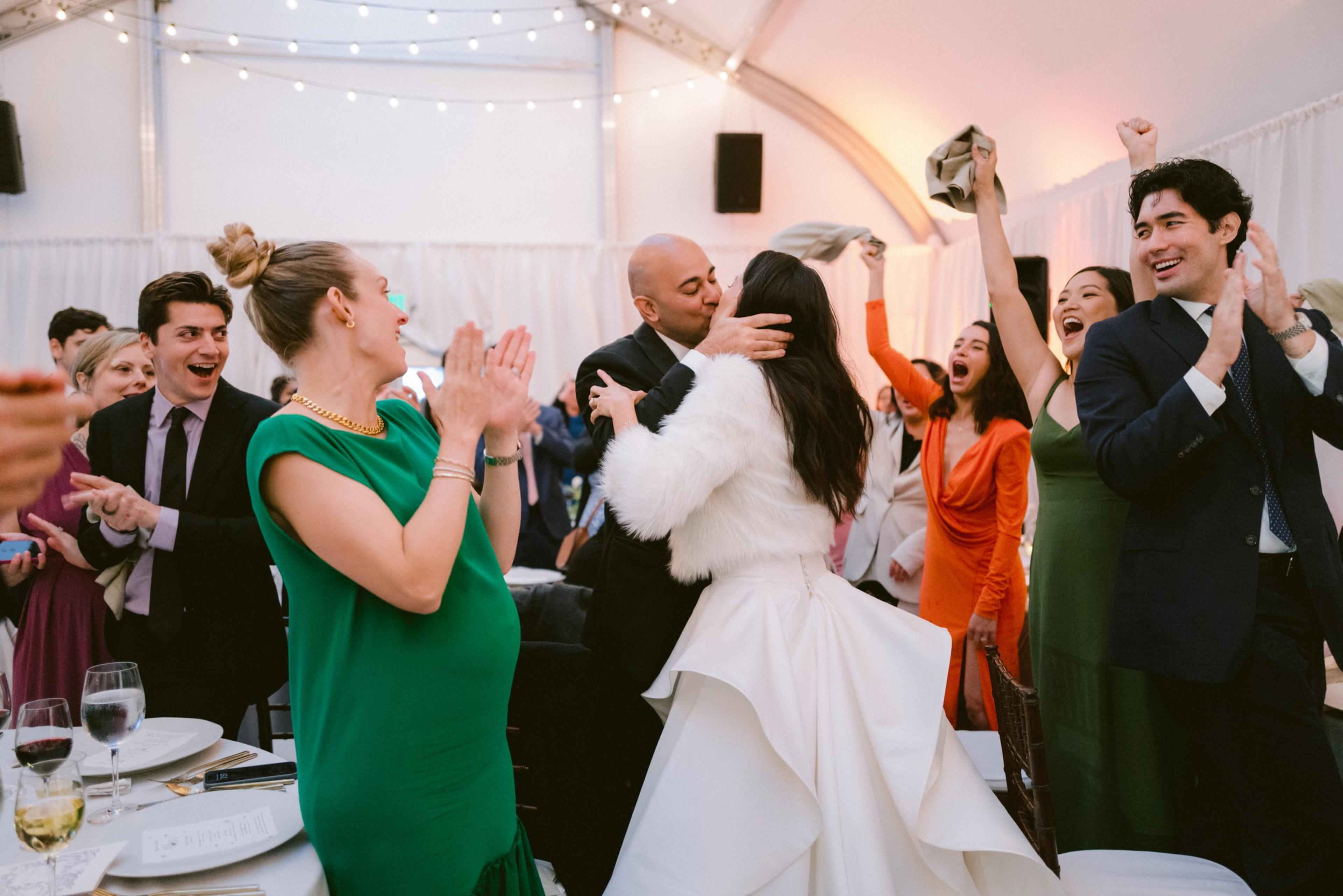 A bride and groom kiss while surrounded by cheering, clapping guests dressed in formal attire at a festive indoor wedding reception.