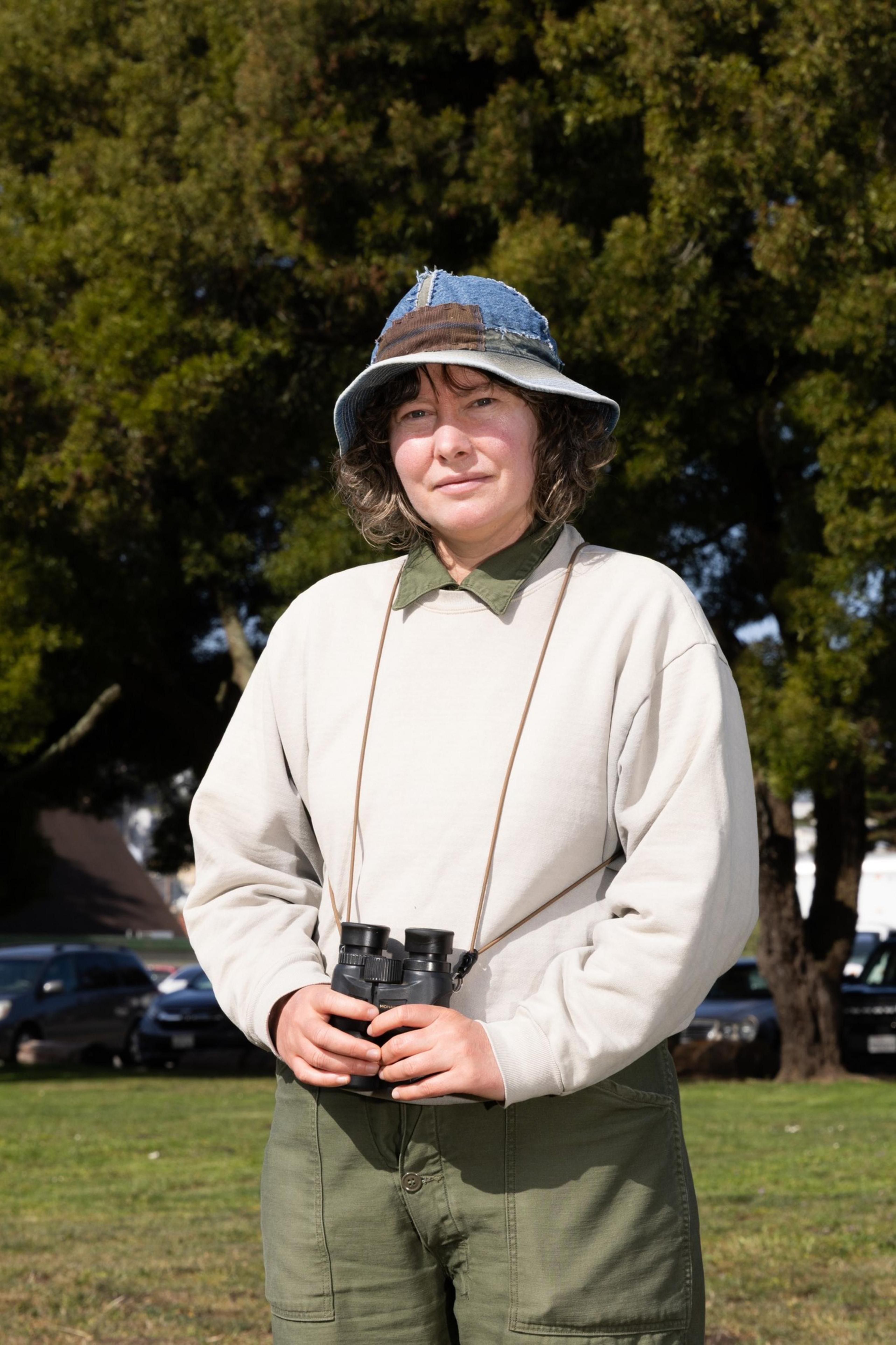 A person stands outdoors, wearing a patched denim bucket hat and a light sweatshirt. They hold binoculars and are in front of trees and parked cars.