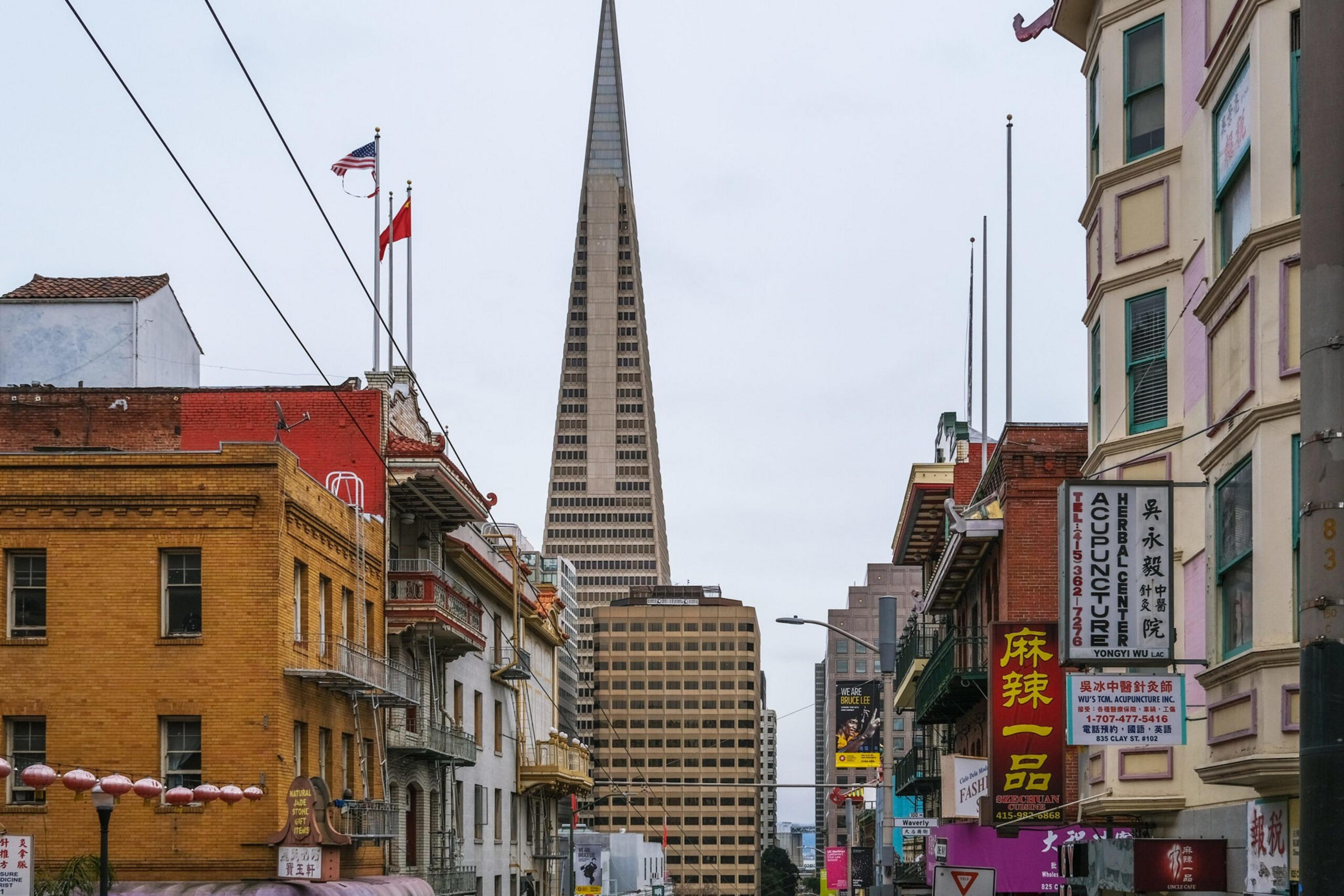 A city street lined with multi-story buildings, featuring the tall, pointed Transamerica Pyramid skyscraper in the background under a gray sky.