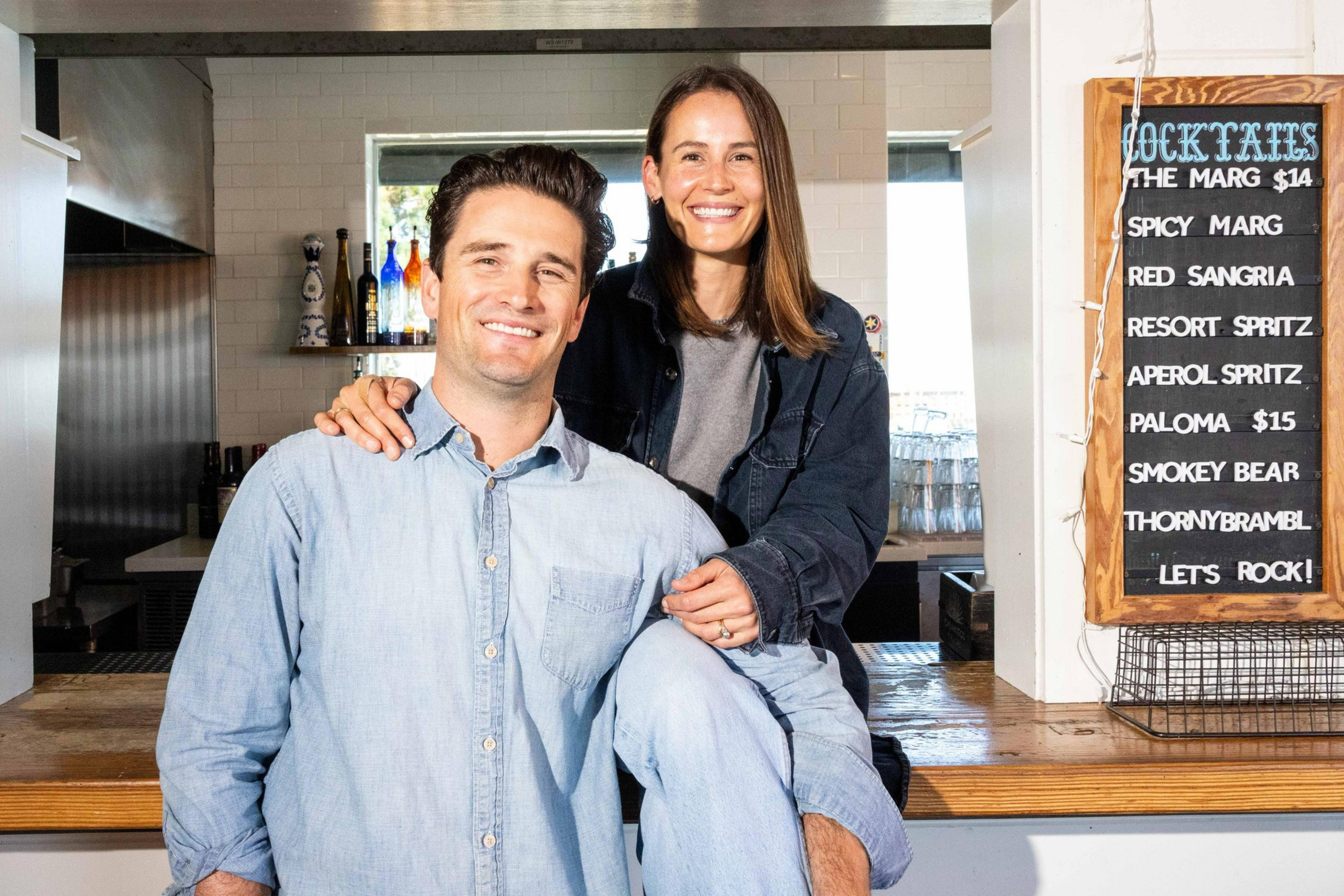 A smiling man and woman pose together indoors by a bar. The woman is sitting on the counter, and a menu board with cocktail names is visible beside them.