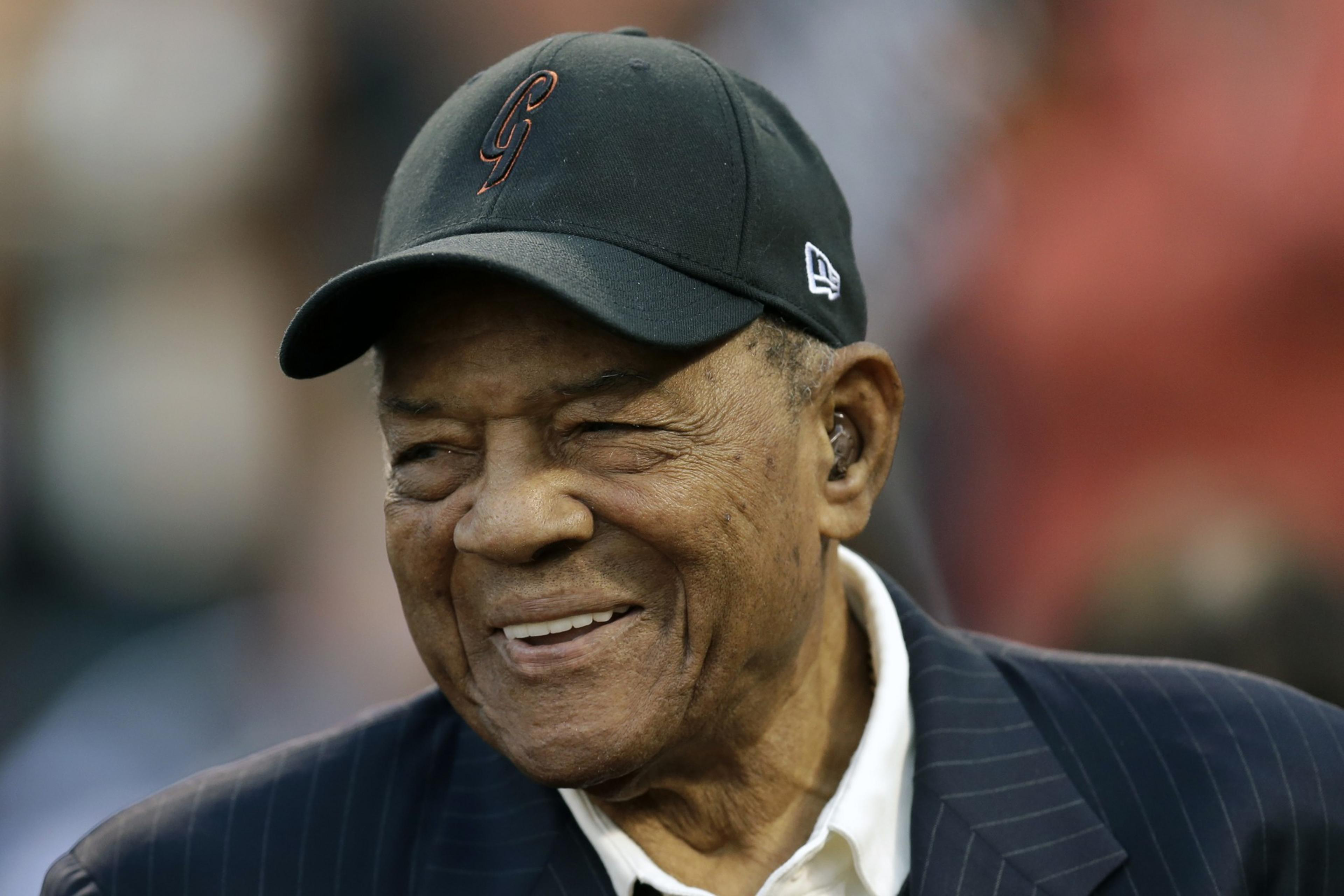 An elderly man with a warm smile is wearing a black baseball cap and a dark striped suit jacket over a white shirt. The background is blurred.