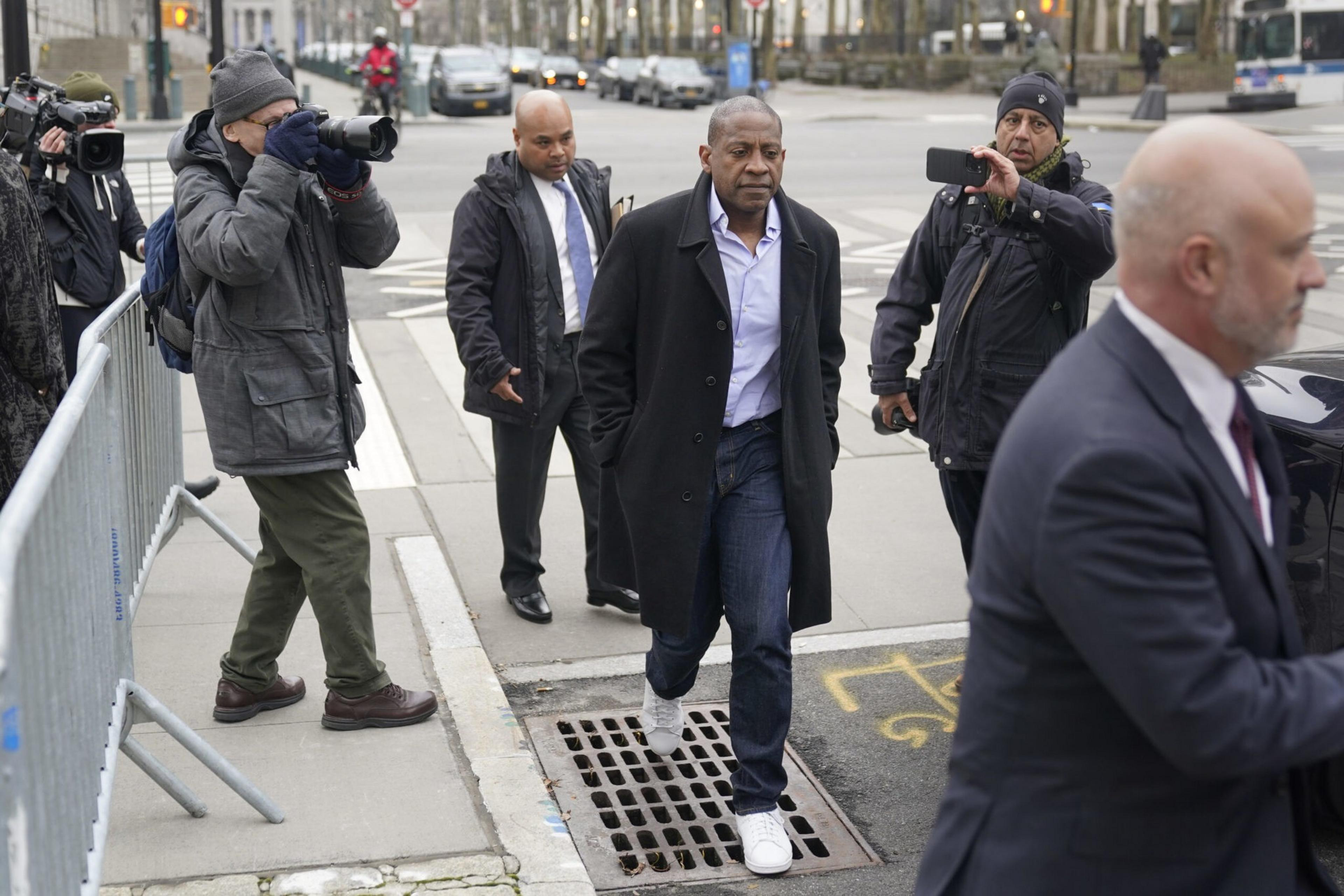 A man in a black coat walks on a sidewalk while photographers and another man in a suit follow him near a street intersection.