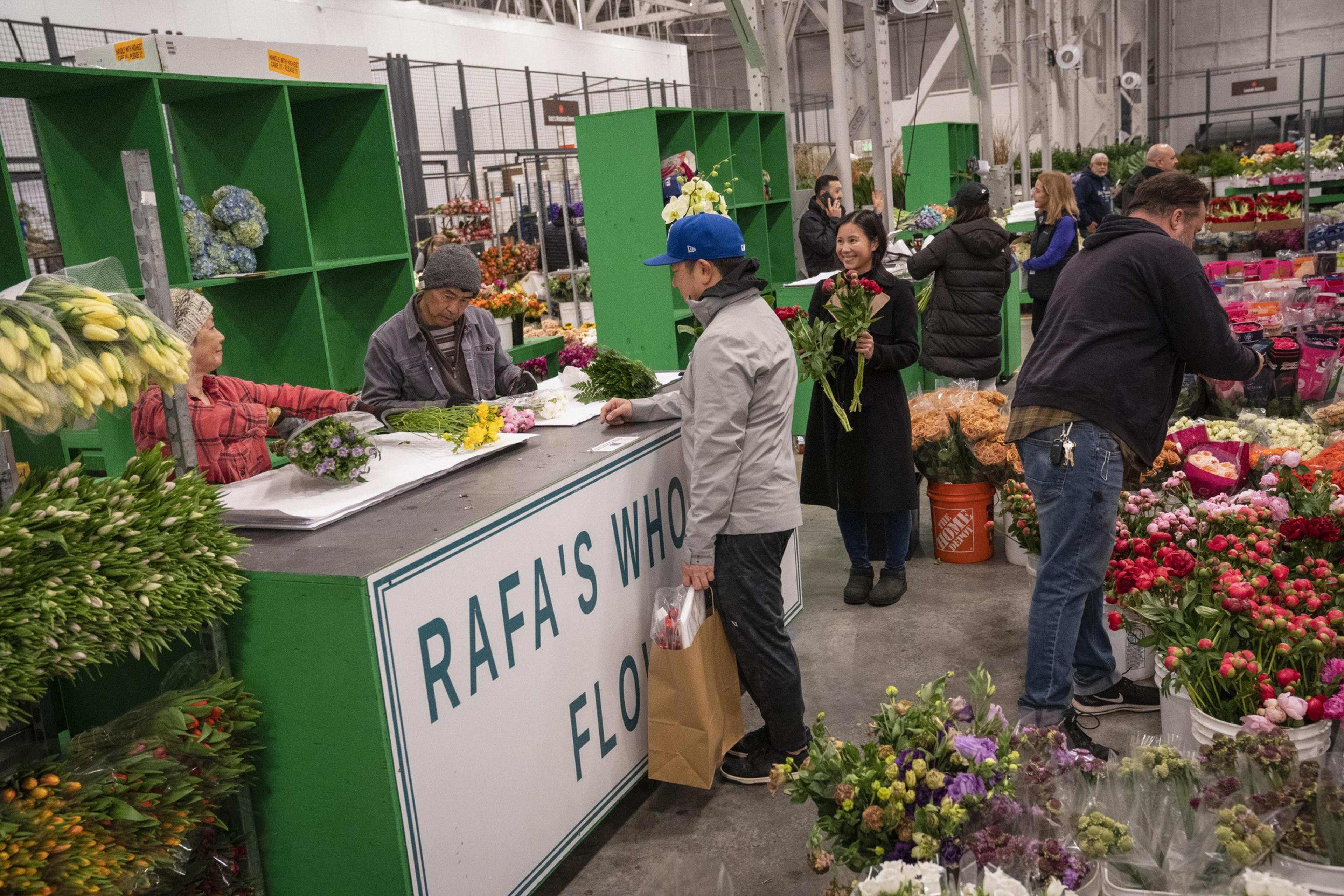 People are buying flowers at a bustling flower market, with vibrant bouquets on display and interactions at a counter labeled &quot;Rafa's Wholesale Flowers.&quot;